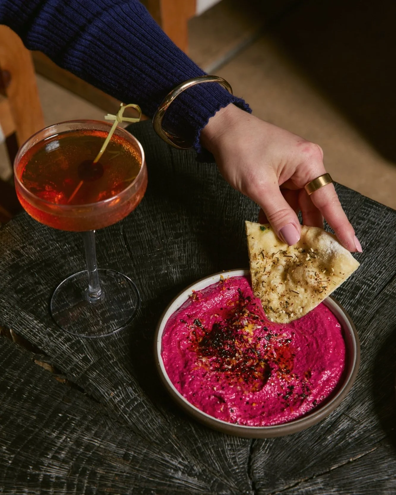 A hand holding a slice of naan bread above a bowl of bright pink beet hummus, with a glass of amber-colored cocktail nearby.