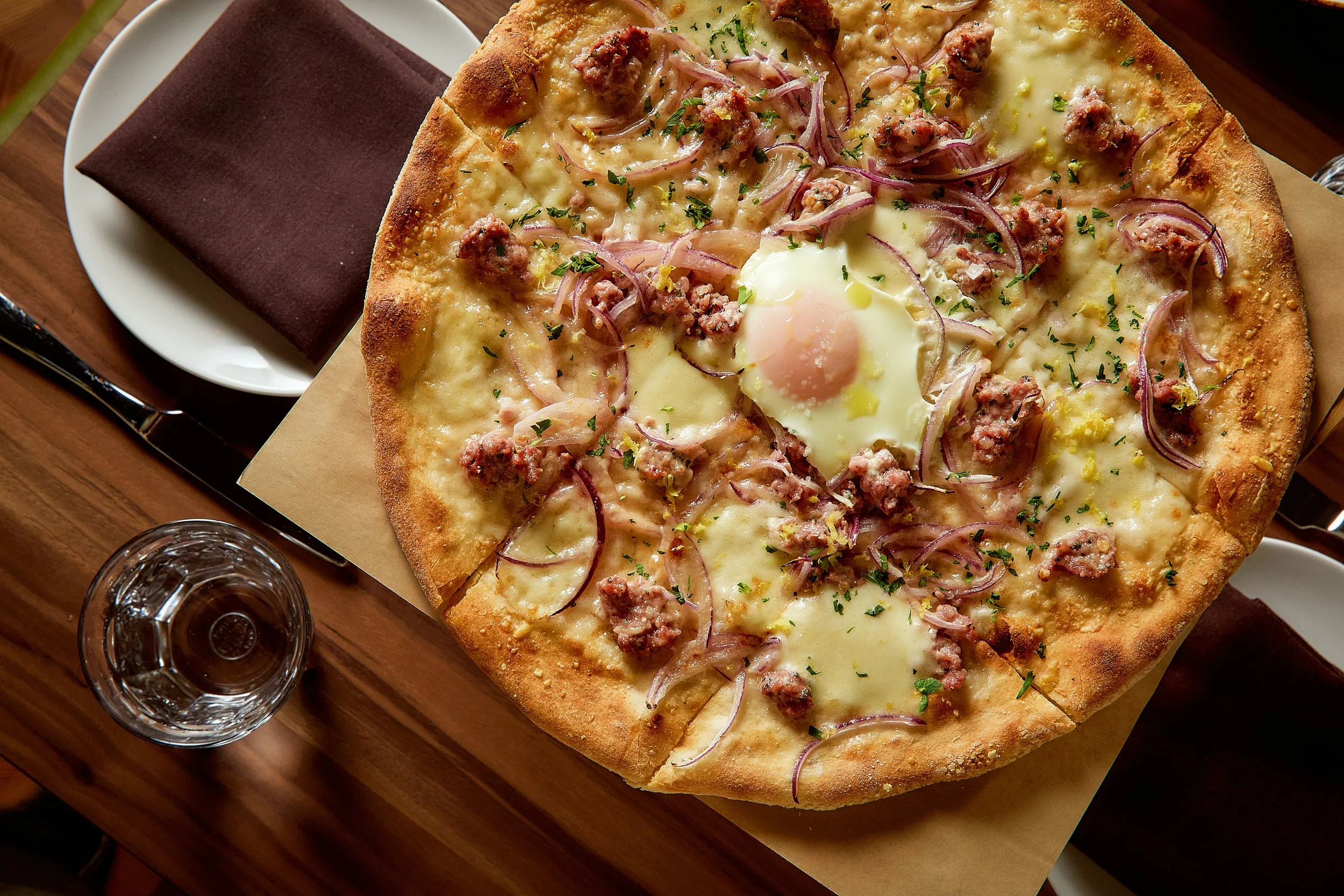 A square pizza topped with cheese, sausage, onions, and herbs, served on a wooden table with a glass of water and a chocolate cake on a white plate.