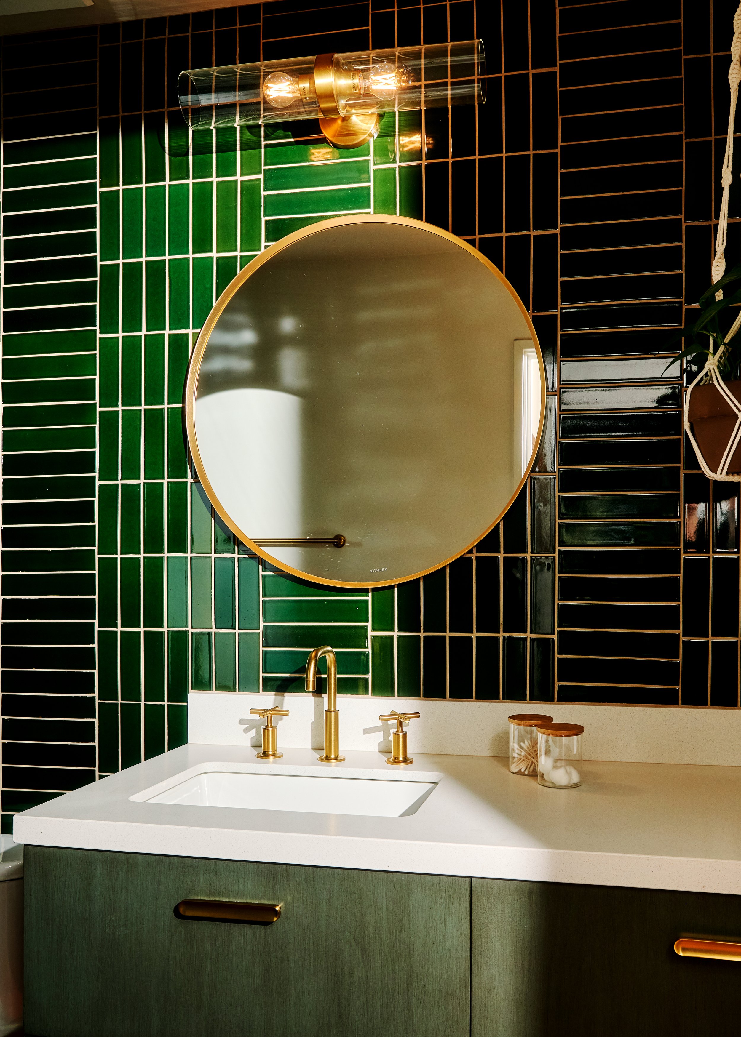 Bathroom vanity with a white countertop, green cabinet, gold faucet, round mirror, green and black tiled wall, decorative containers, and a modern wall light fixture.