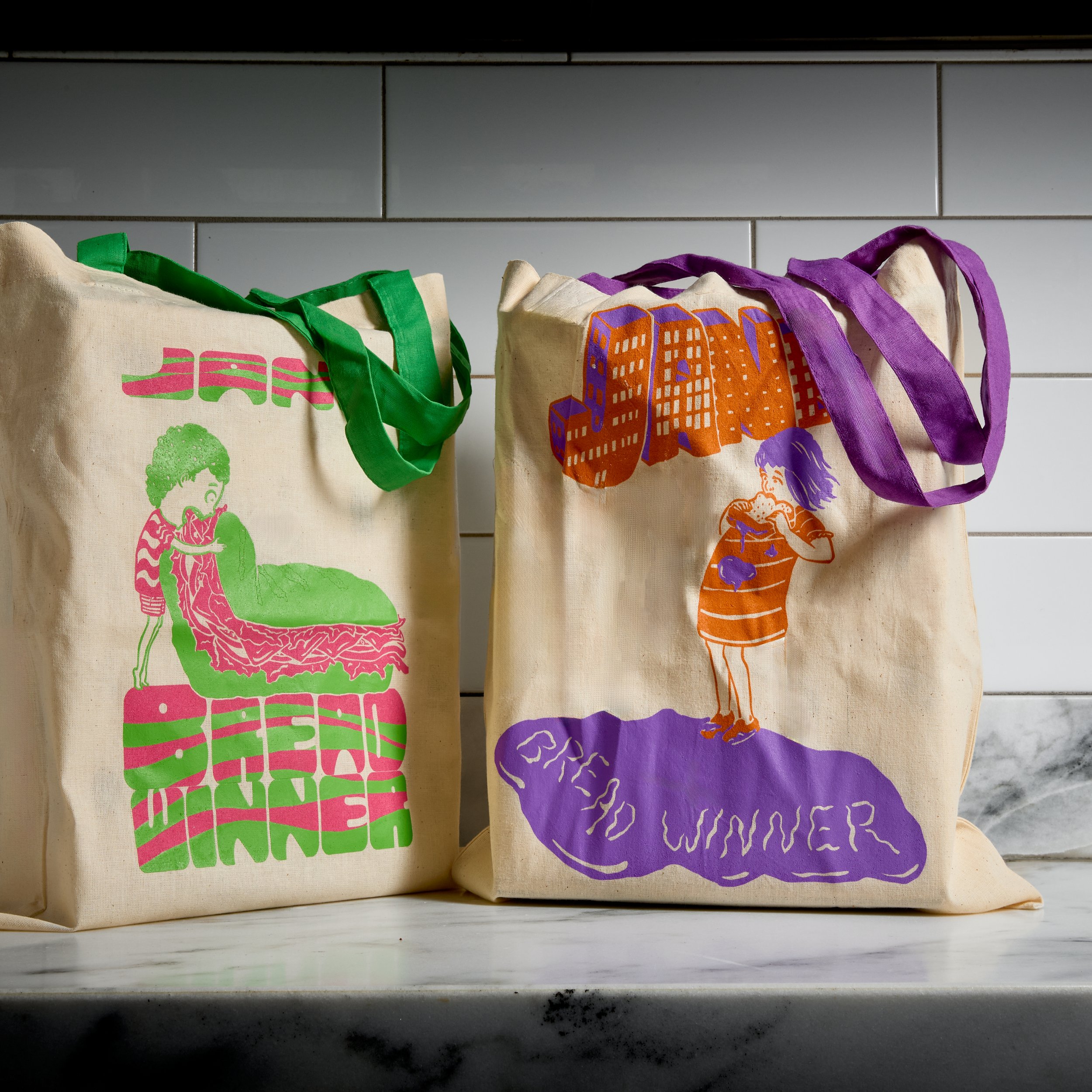 Two canvas tote bags with colorful illustrations and texts on a white marble surface, against a white tiled wall. The left bag has green handles and a design with a woman sitting on a couch and the text 'JANE'S DINNER'. The right bag has purple handl