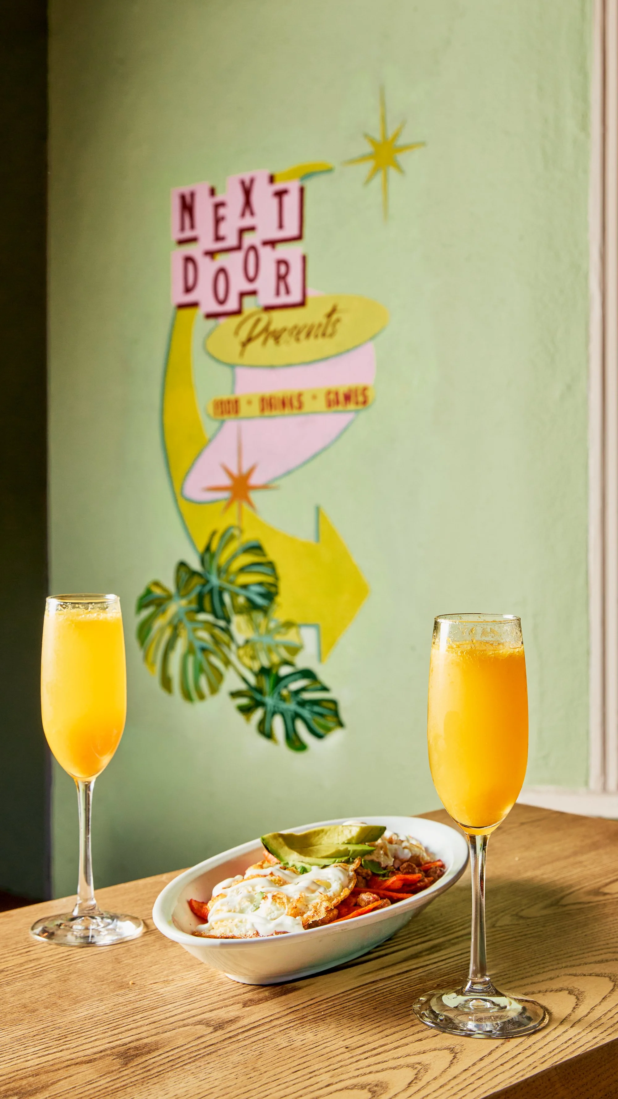 A bowl of chili topped with sour cream and avocado slices, flanked by two glasses of orange mimosa on a wooden table, with a colorful retro sign for Next Door Presents on a green wall in the background.