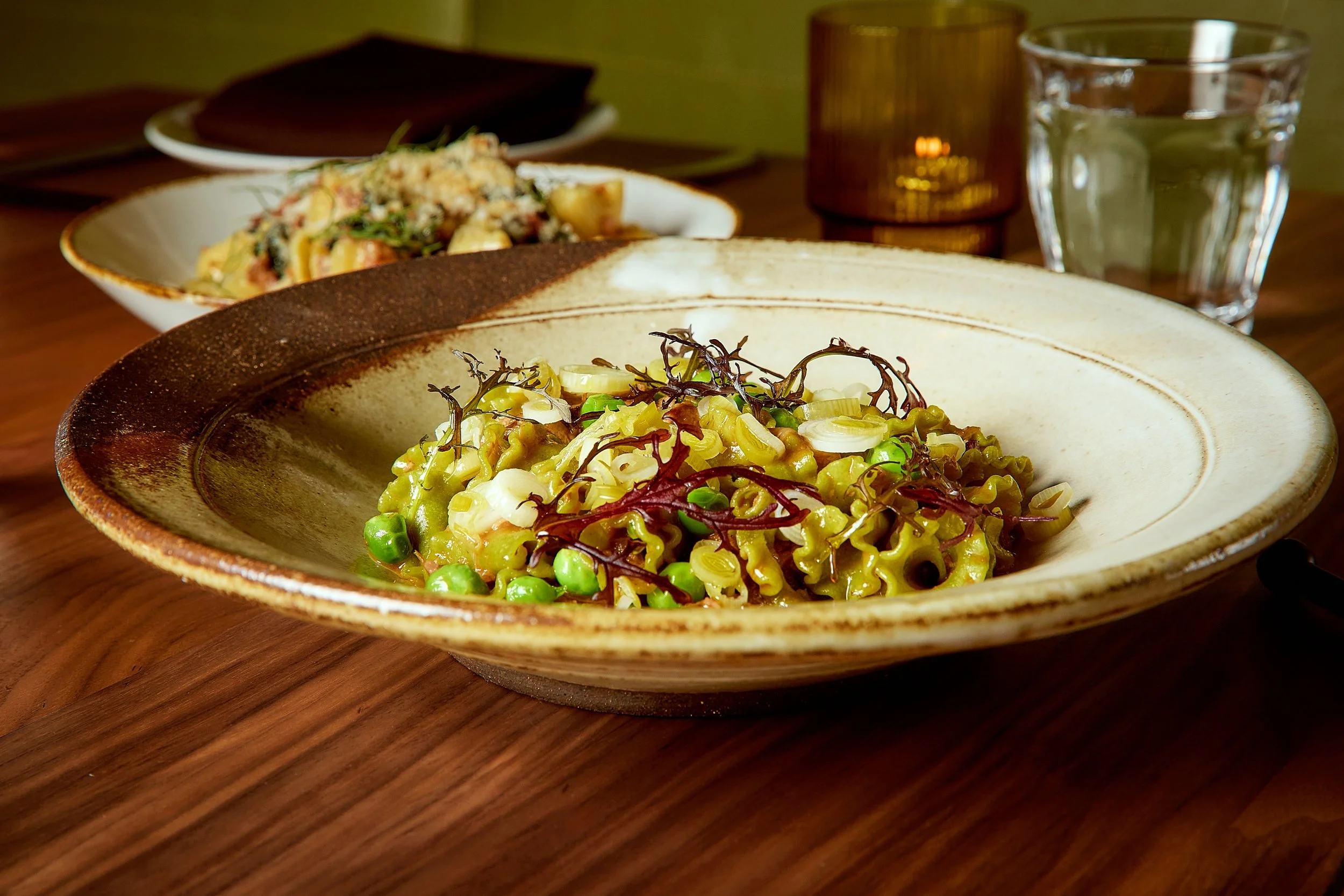 A bowl of cooked pasta with green peas, shredded vegetables, and garnished with herbs on a wooden table, with another dish and glasses in the background.