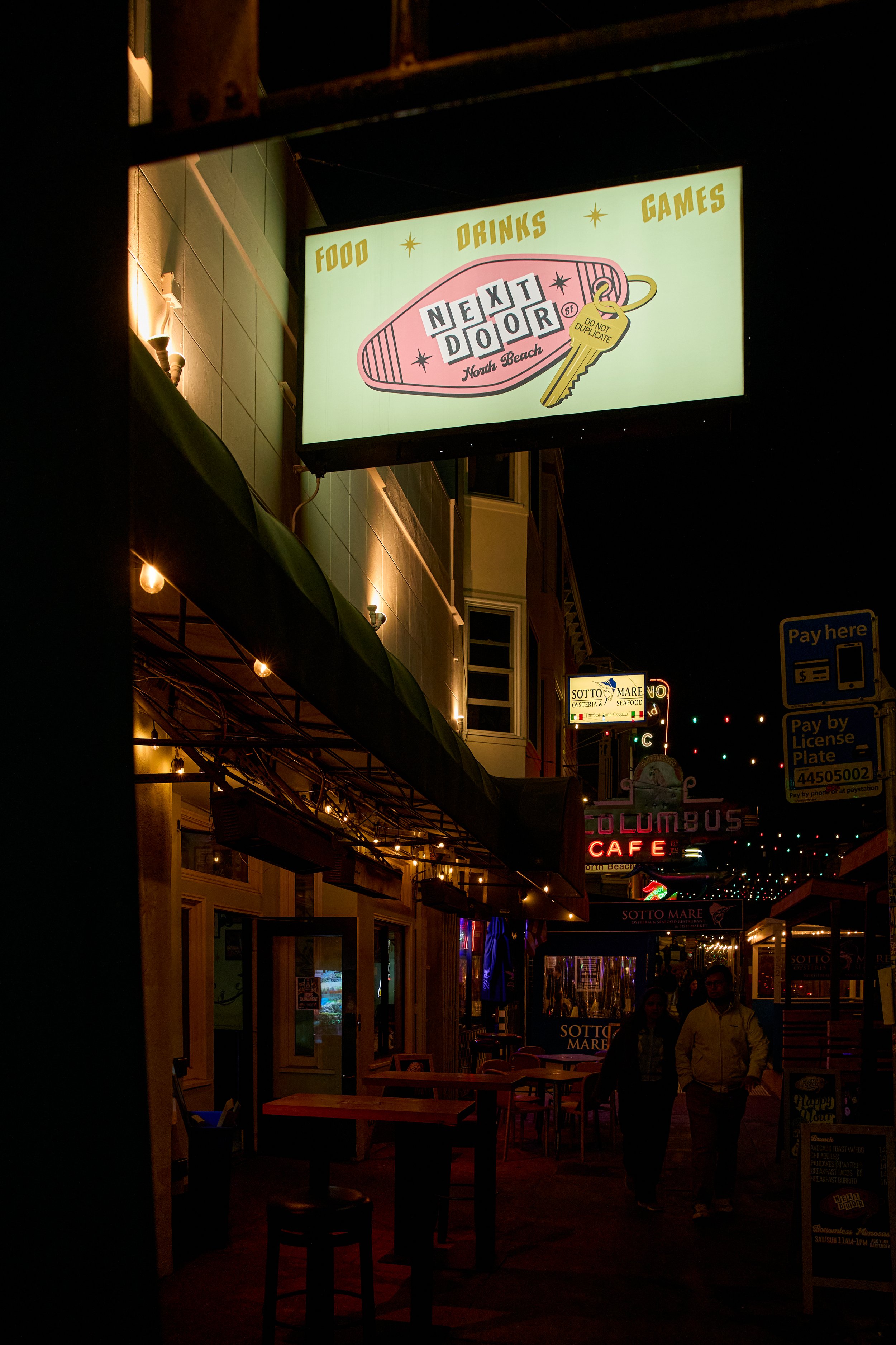 Night view of a street with illuminated signs, including a large sign for Next Door North Beach, with food, drinks, and games, and smaller signs for Sotto Mare and Columbus Cafe, with outdoor seating and pedestrians walking.