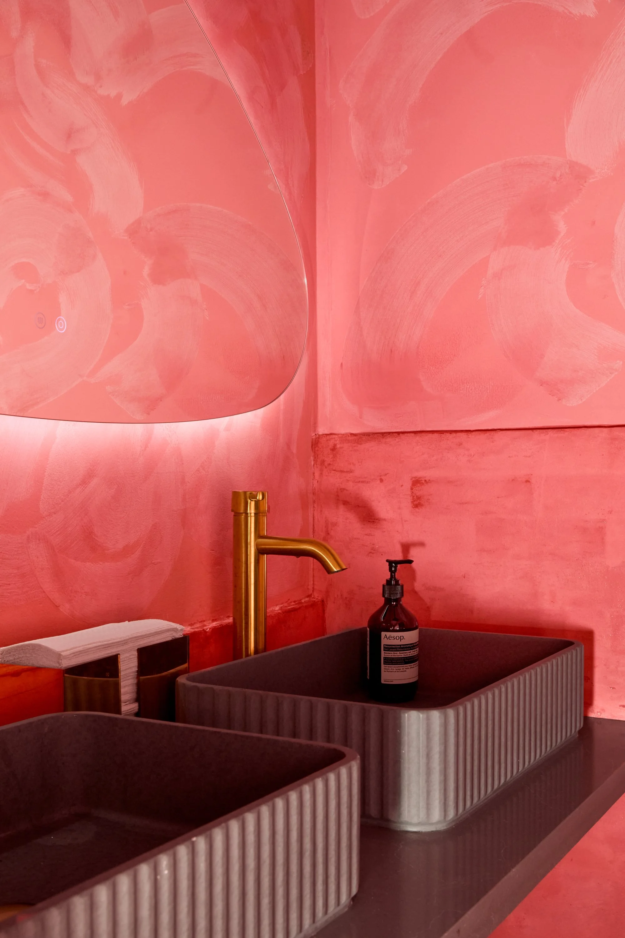 Two rectangular sinks with a ribbed design, one dark and one light, with a gold faucet in a pink-painted room. A bottle of Aesop hand soap is in one sink, and tissue paper is on a holder nearby.