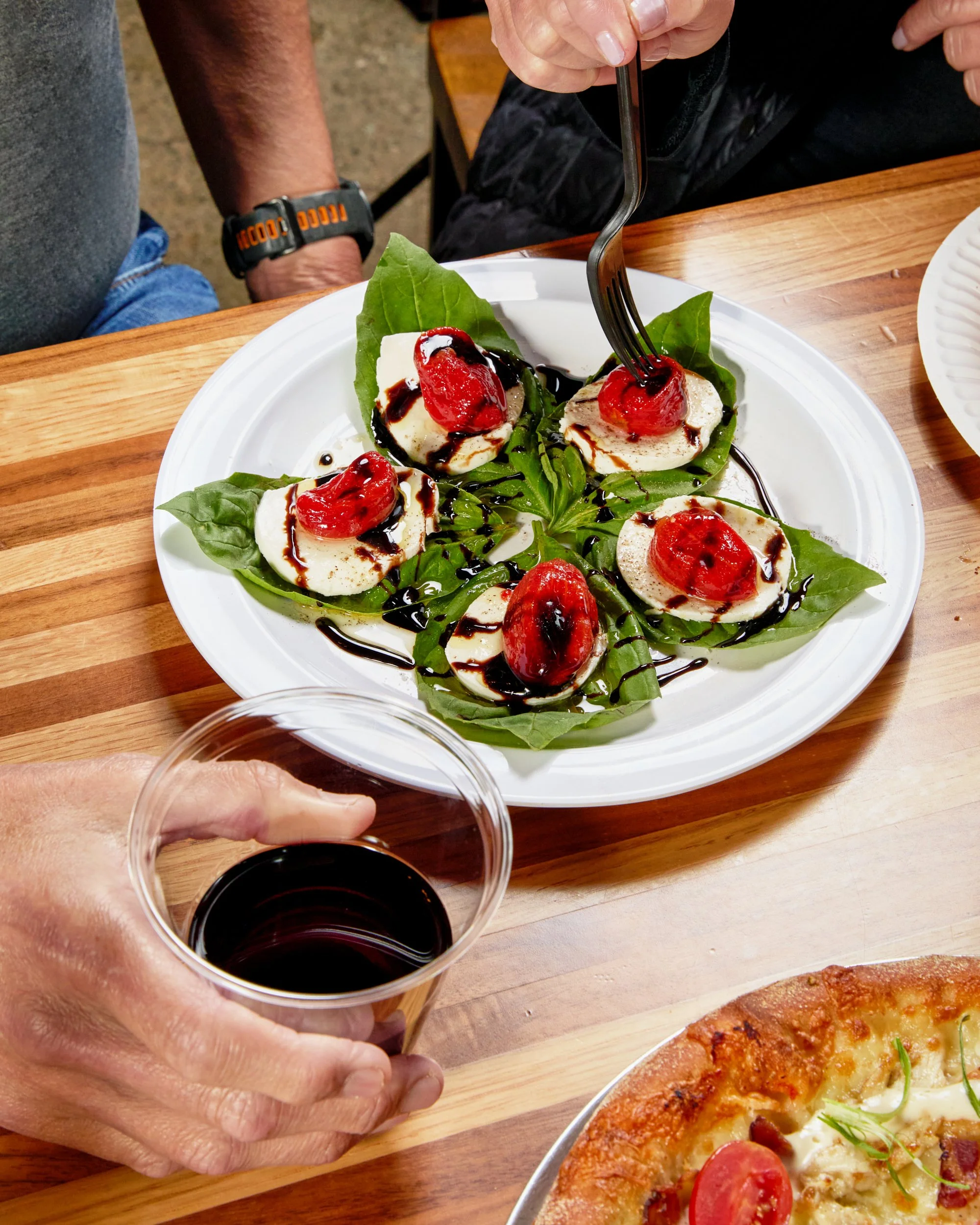 A person holding a glass of red wine, with a pizza and a plate of mozzarella cheese salad topped with cherry tomatoes, basil, and balsamic glaze on a wooden table.