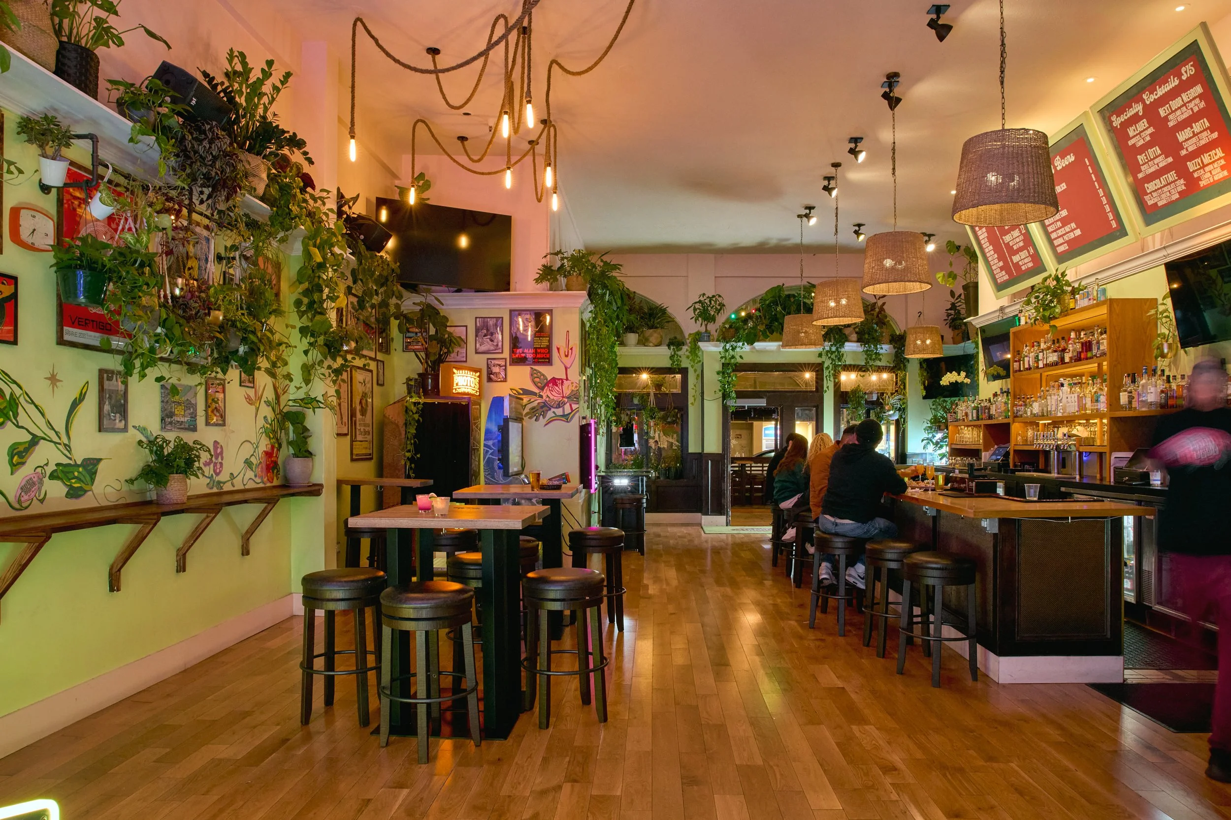 Interior of a cozy bar with wooden flooring, green walls decorated with plants and framed pictures, and a bar counter with stools. Pendant and string lights hang from the ceiling, and a group of people sit at the bar.