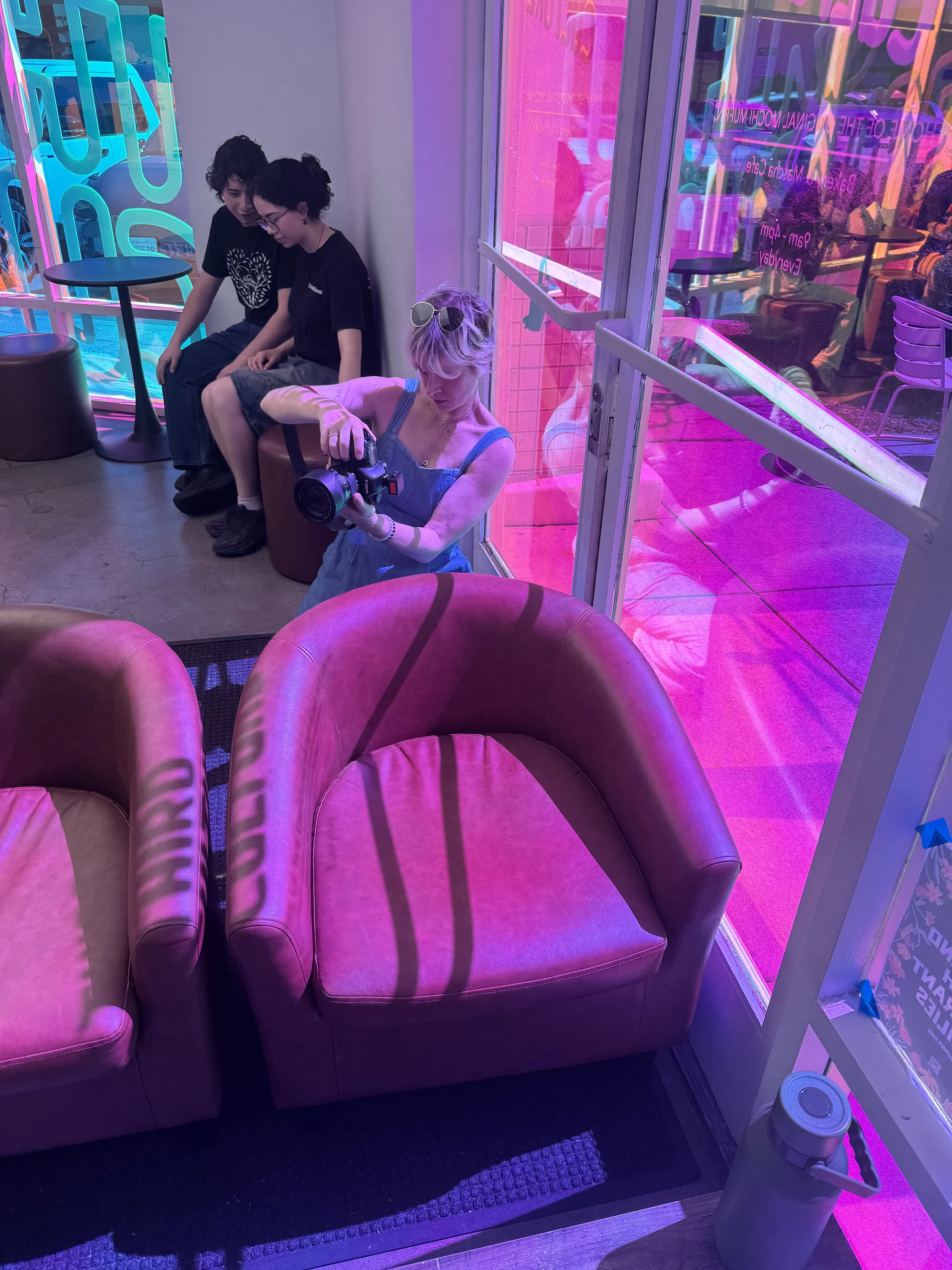 A group of three people sitting on couches in front of a brightly lit, colorful neon sign and windows with neon lights. One person with blonde hair is using a camera, two others with dark hair are sitting together, looking downward.