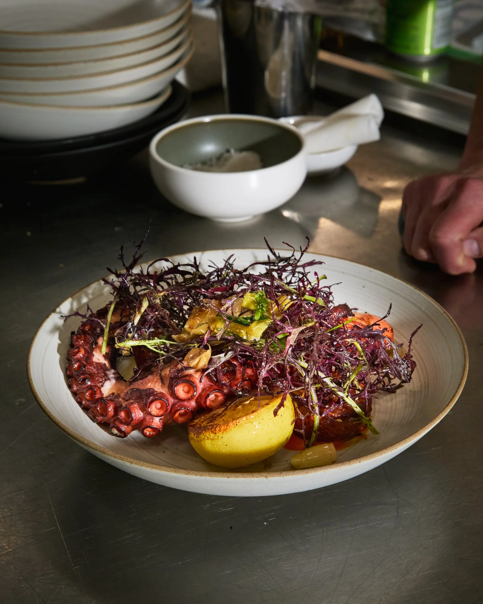Plate of grilled octopus with purple microgreens and lemon wedge on a gray ceramic dish, in a restaurant kitchen setting.