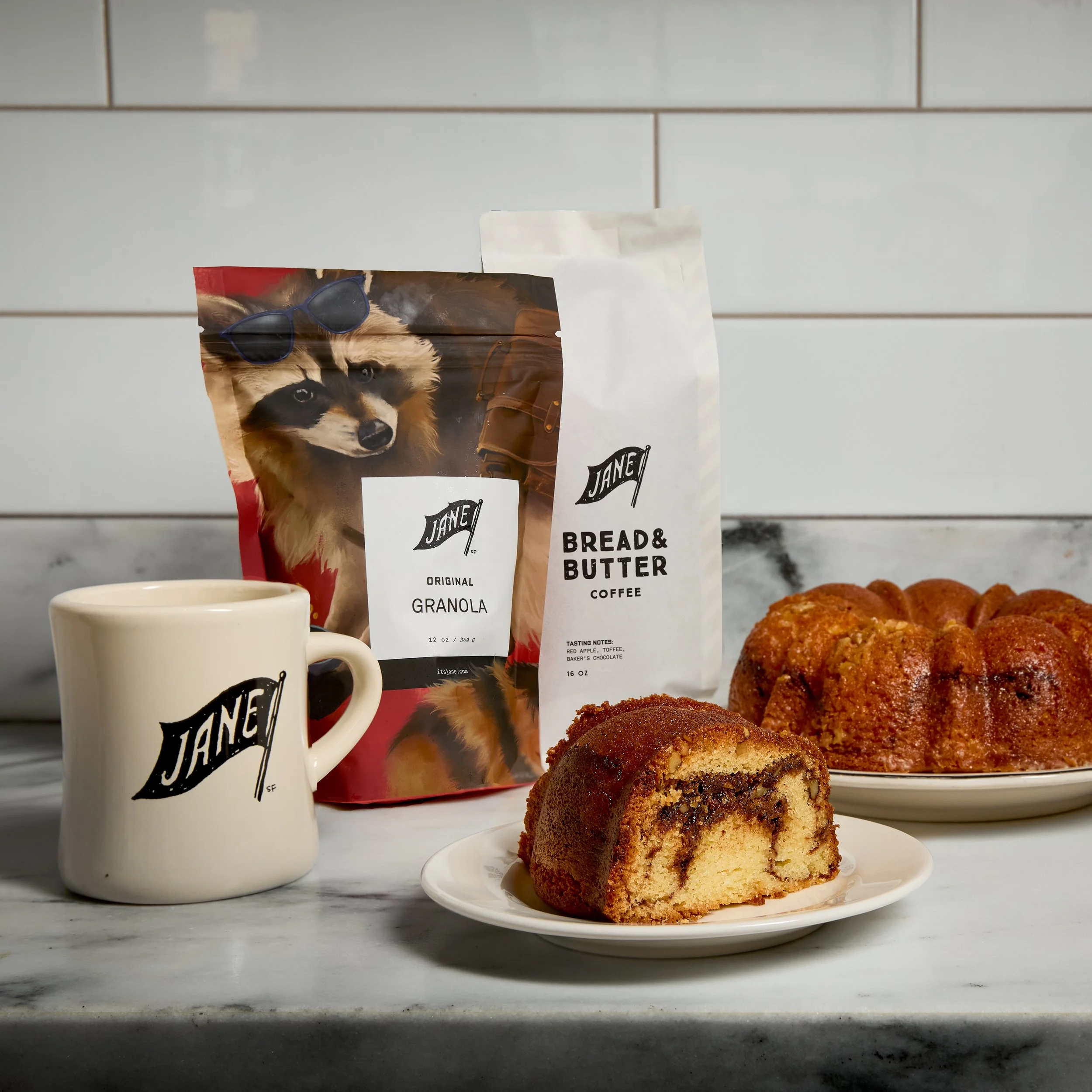 Coffee mug, bag of Jane's granola, bag of Jane's bread and butter coffee, slice of marble cake on a plate, and a bundt cake on a plate, all arranged on a marble surface countertop with white tiled wall background.