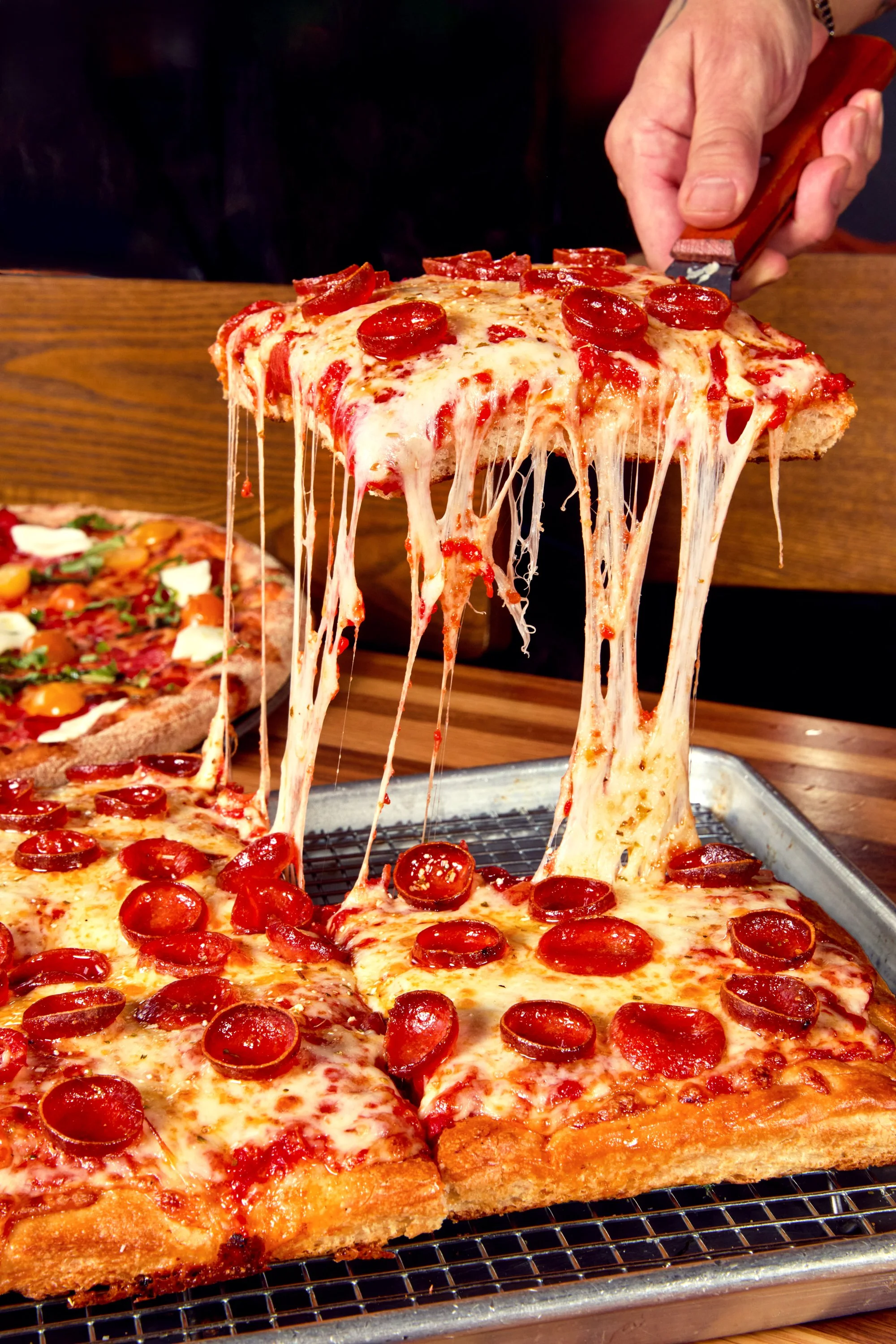 Close-up of a slice of pepperoni pizza with melted cheese being lifted, stretching as the slice is removed from the pizza on a baking sheet.