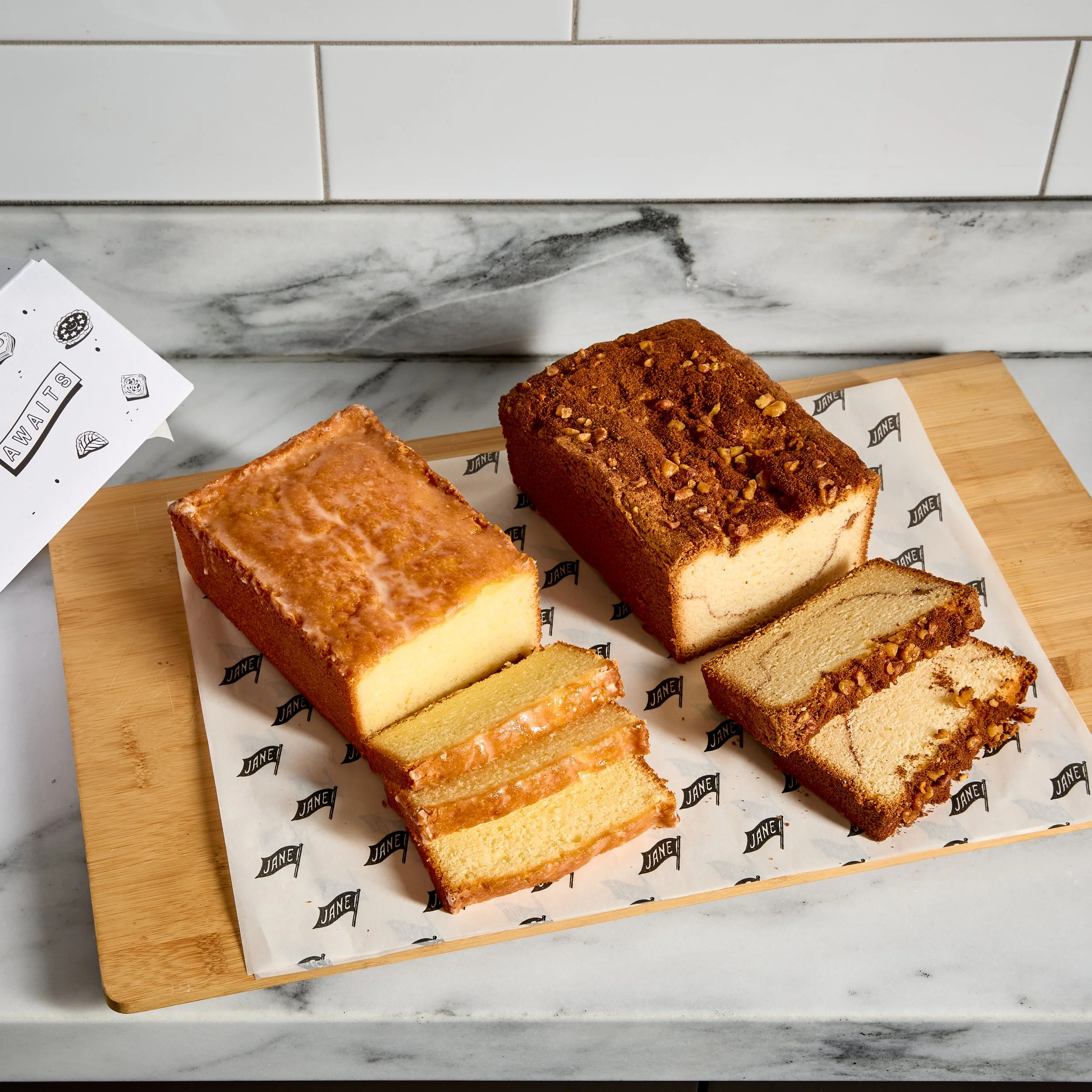 Assorted slices of vanilla and chocolate marble cakes on a wooden tray covered with branded paper, with a marble and tile background.