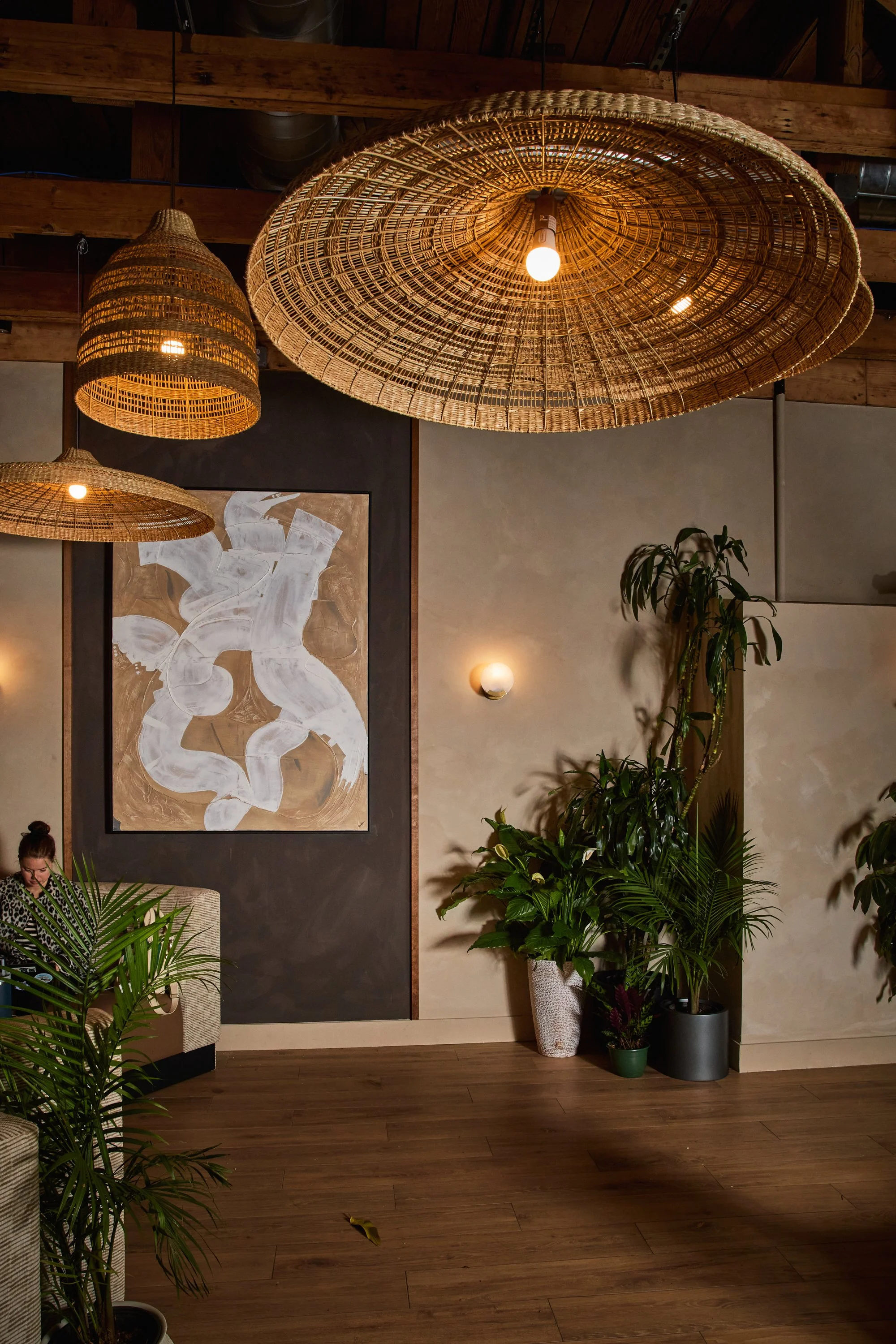 Indoor space with wooden floors, large woven pendant lights, abstract art on wall, and lush potted plants.