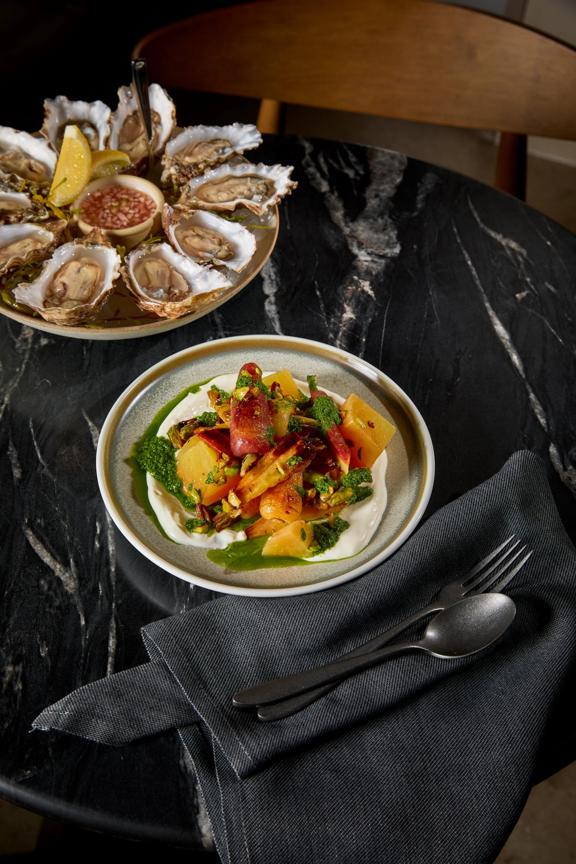 A black marble table set with a bowl of roasted vegetables topped with herbs and a plate of raw oysters on the half shell with lemon wedges and cocktail sauce.
