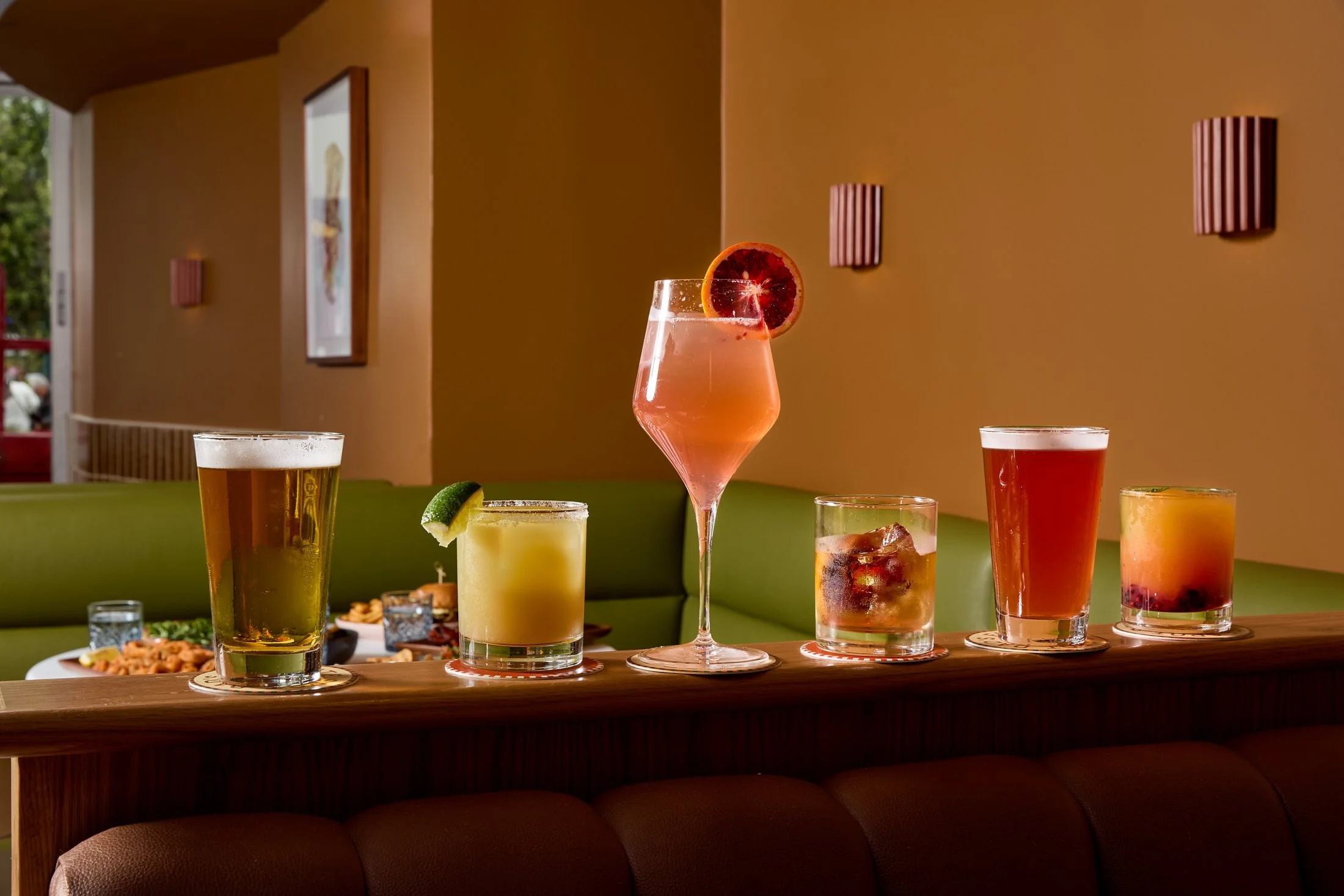 A variety of colorful cocktails on a wooden bar counter in a restaurant with green seating and decor on a beige wall.