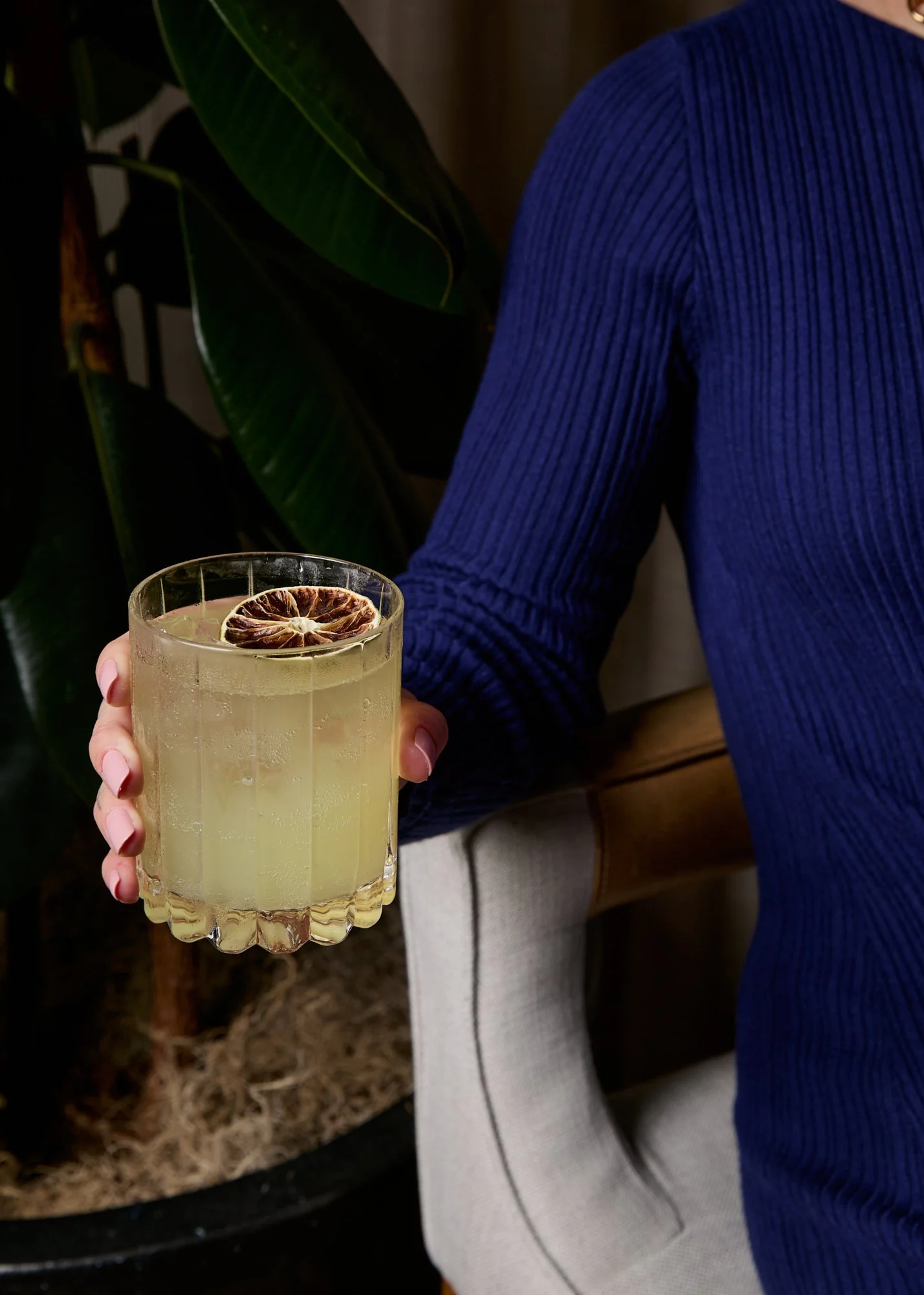 A person holding a glass of light-colored cocktail garnished with a dried citrus slice, wearing a blue ribbed sweater.