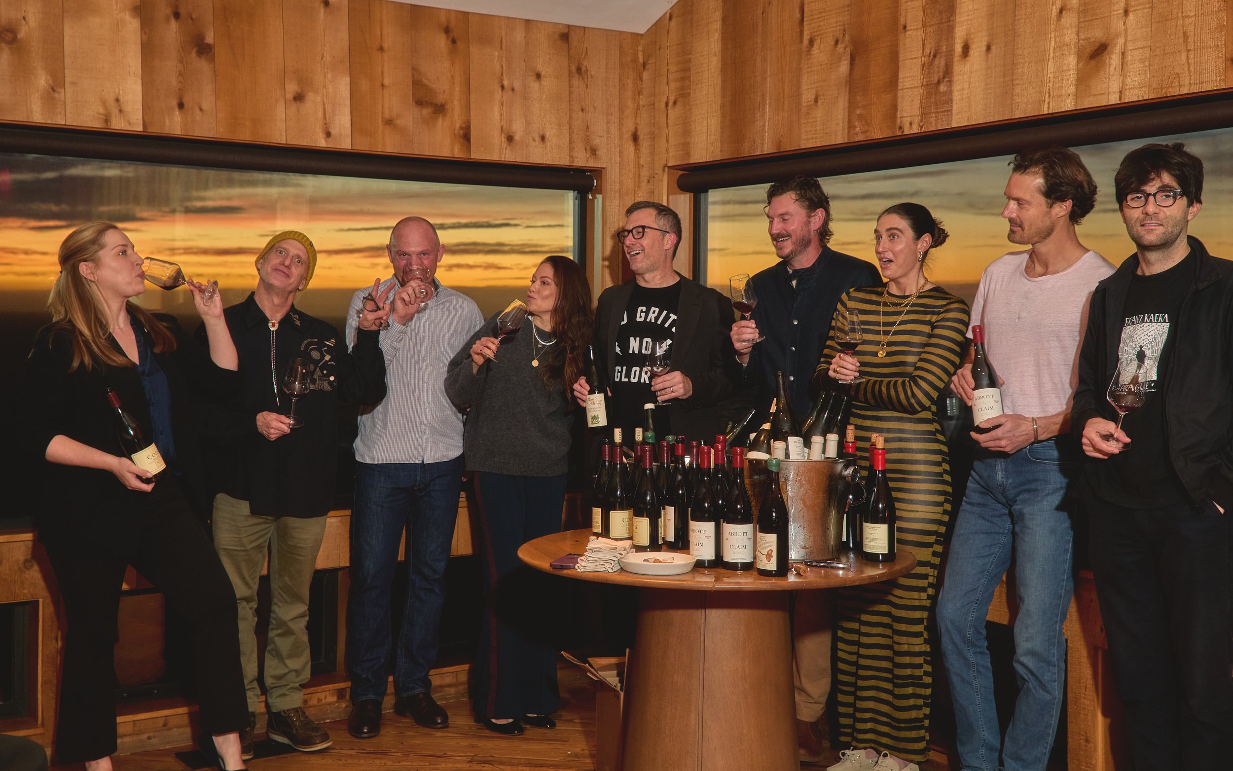 A group of ten people standing together indoors during sunset, holding glasses of wine in front of windows showing a sunset sky, with a table of wine bottles in front.