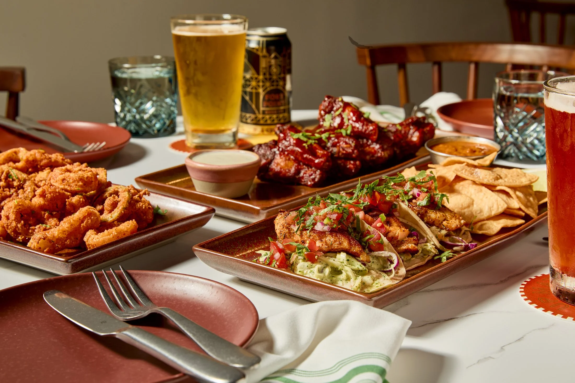 A table filled with Mexican dishes including fried chicken, tacos with meat and vegetables, and grilled meat, along with beverages such as beer and water, set with plates, napkins, and utensils.
