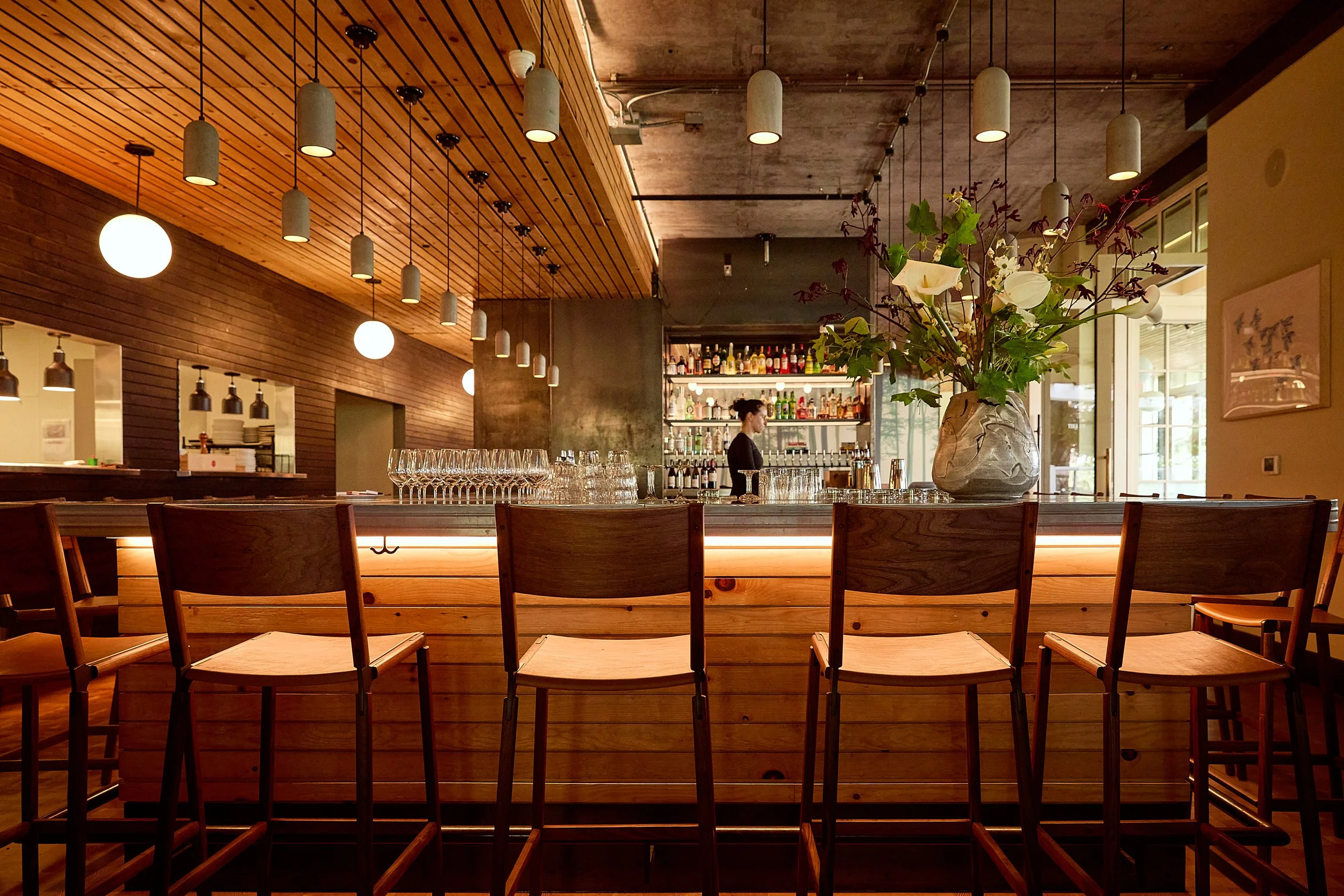 A modern restaurant interior with a wooden bar counter, empty chairs, a large flower arrangement, and a bartender in the background, illuminated by hanging lights.