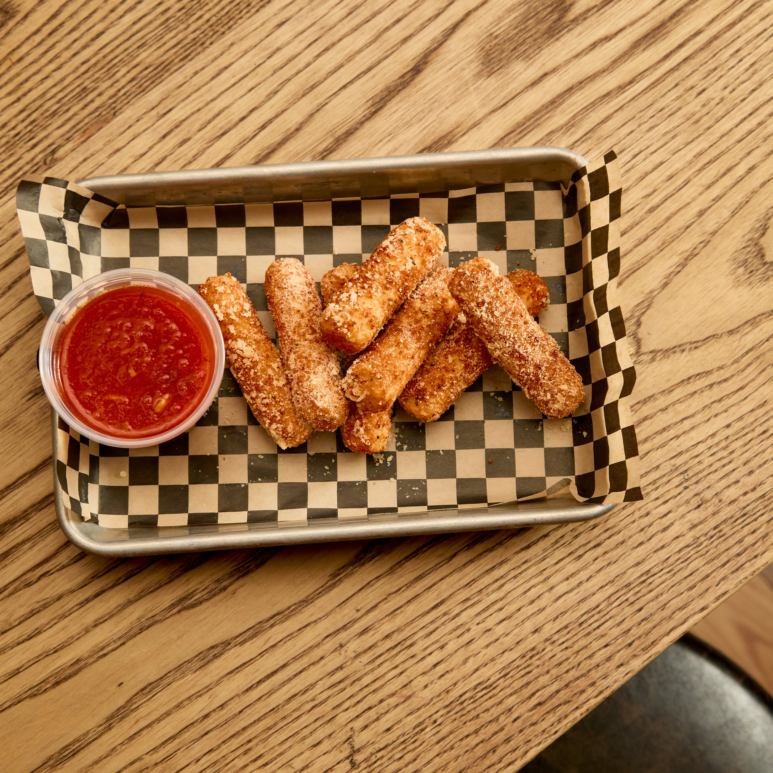 Breaded mozzarella sticks with marinara sauce on a checkered paper-lined metal tray on a wooden table.