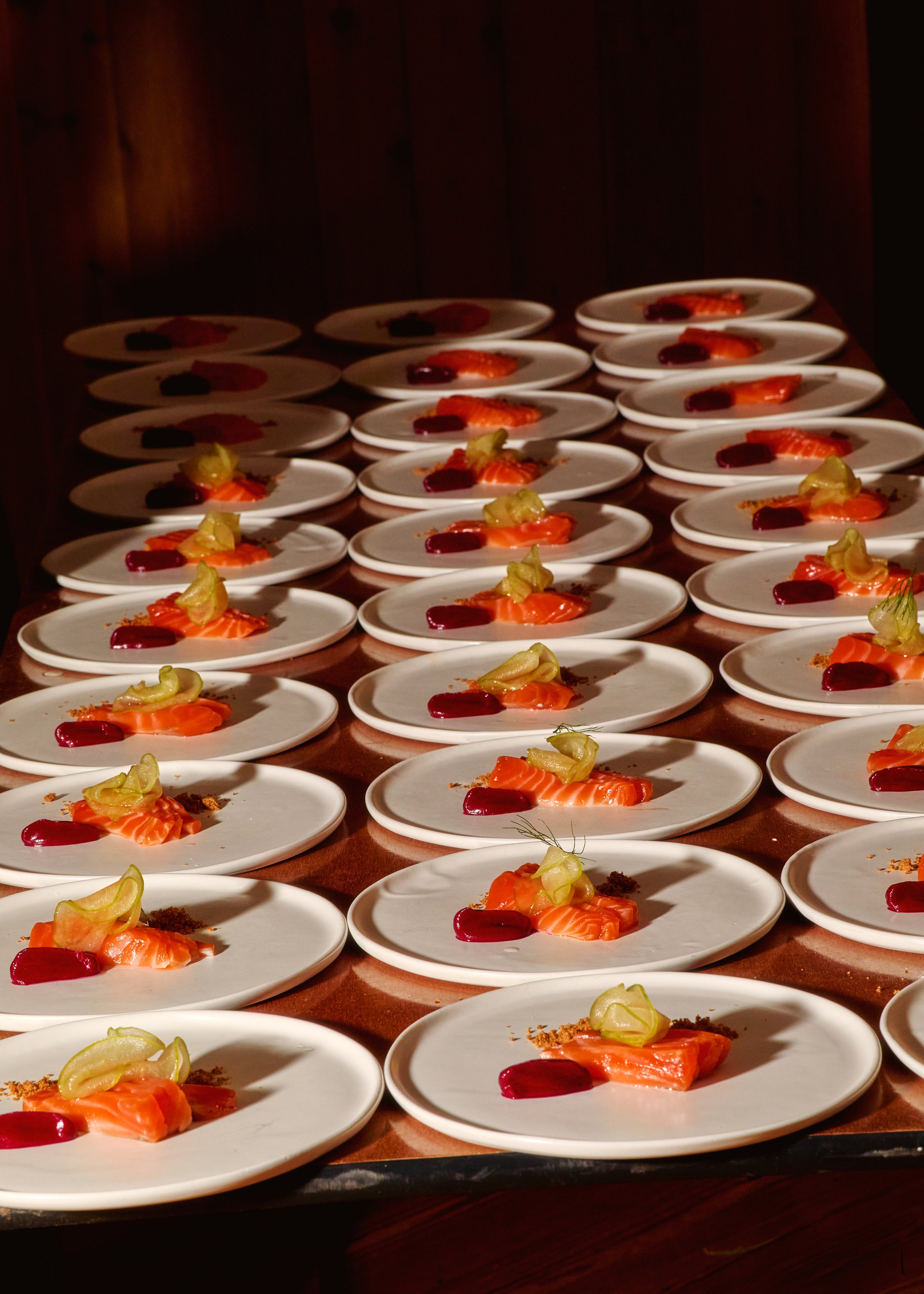 Multiple white plates arranged in rows on a wooden table, each plate holding slices of salmon garnished with pickles and a dark red sauce.