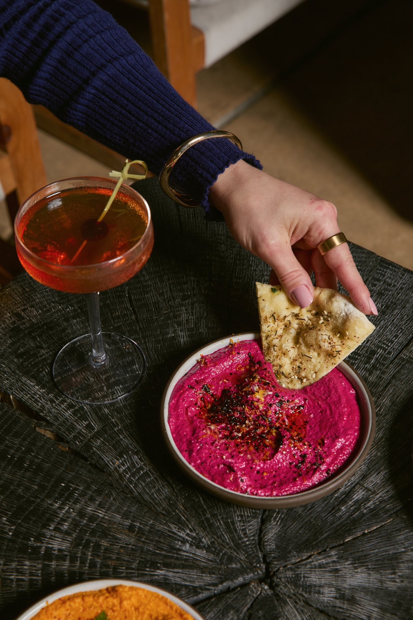 A person wearing a gold bracelet and a gold ring is reaching for a slice of naan bread from a bowl of pink hummus on a black textured table, with a cocktail glass filled with a red drink nearby.