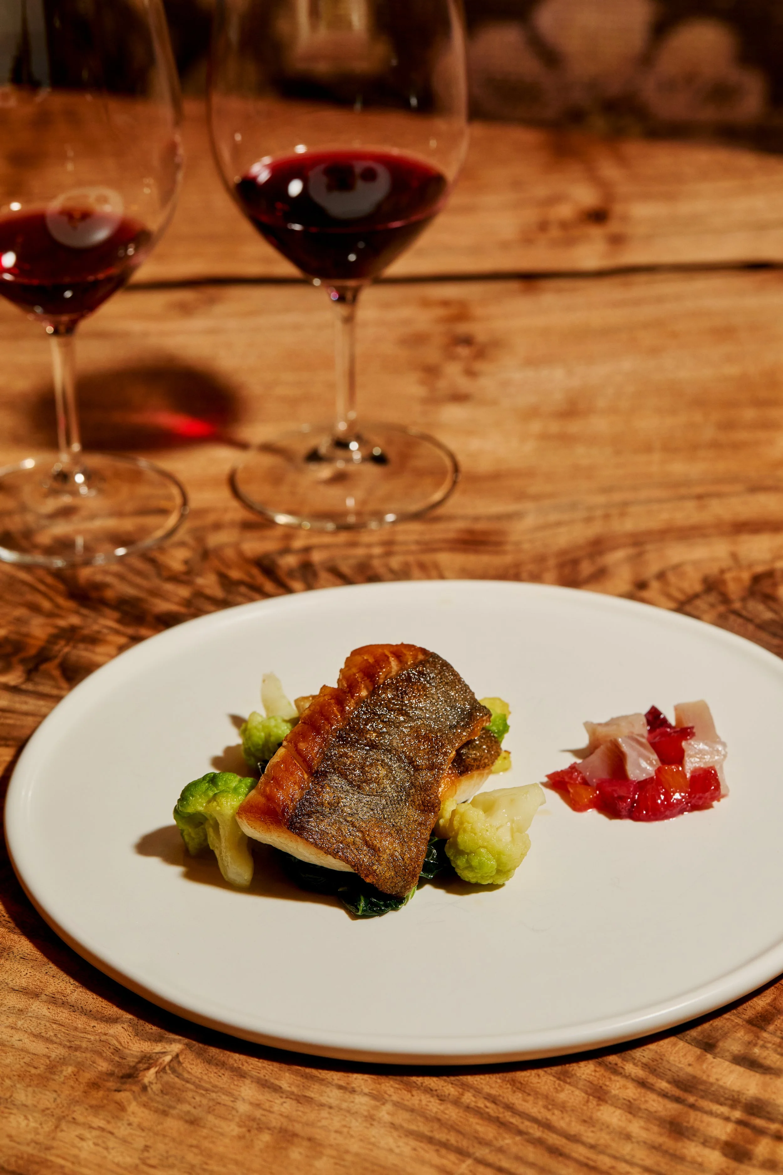 A plated seared fish fillet with vegetables, served alongside diced cold cuts on a white plate, with two glasses of red wine on a wooden table.