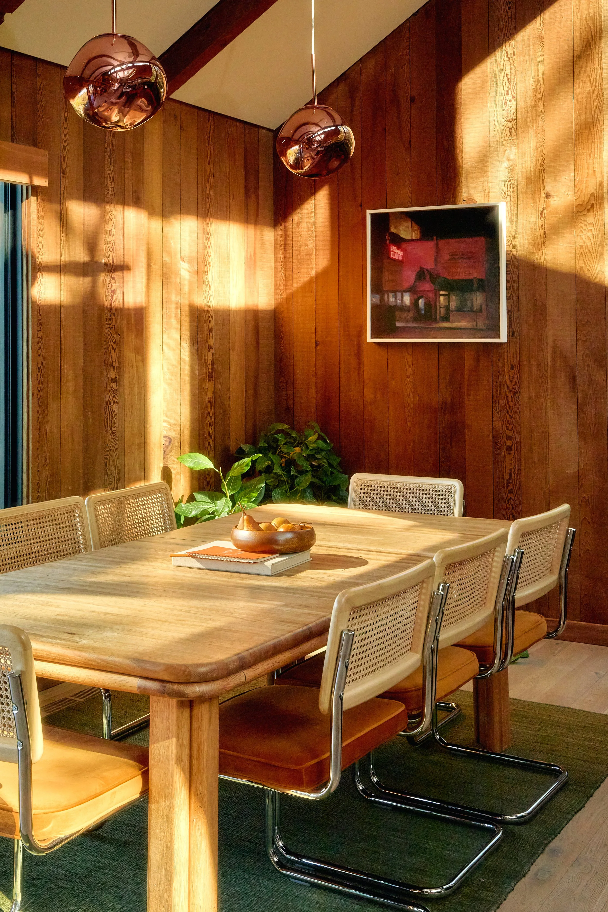 A wooden dining table with six cream-colored chairs, a bowl of apples, and a closed notebook. A green potted plant is beside the table. The background features a wooden paneled wall, a framed painting, and two hanging copper pendant lights. Sunlight 