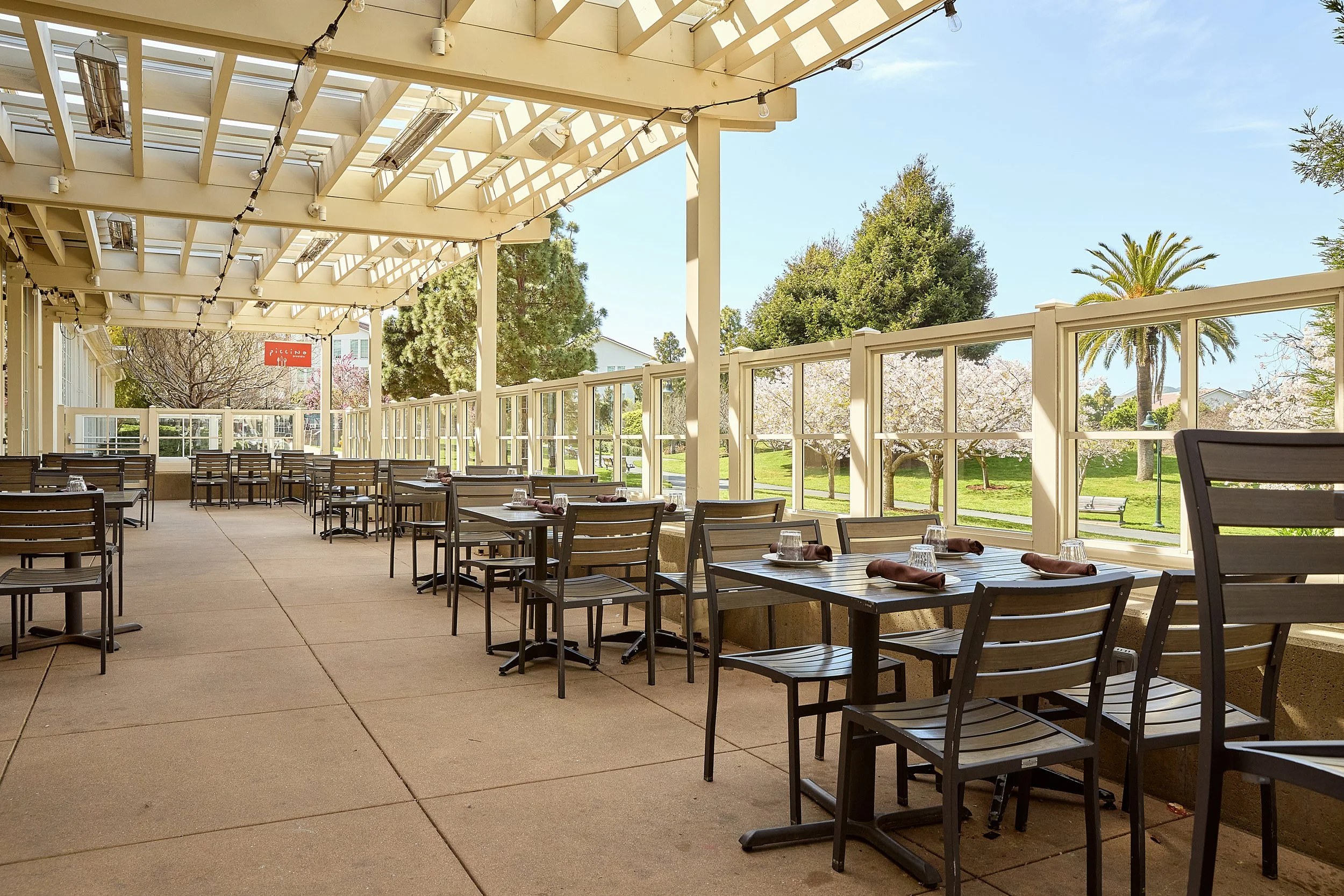 Empty outdoor patio with wooden tables and chairs, string lights hanging overhead, surrounded by trees and a blue sky.