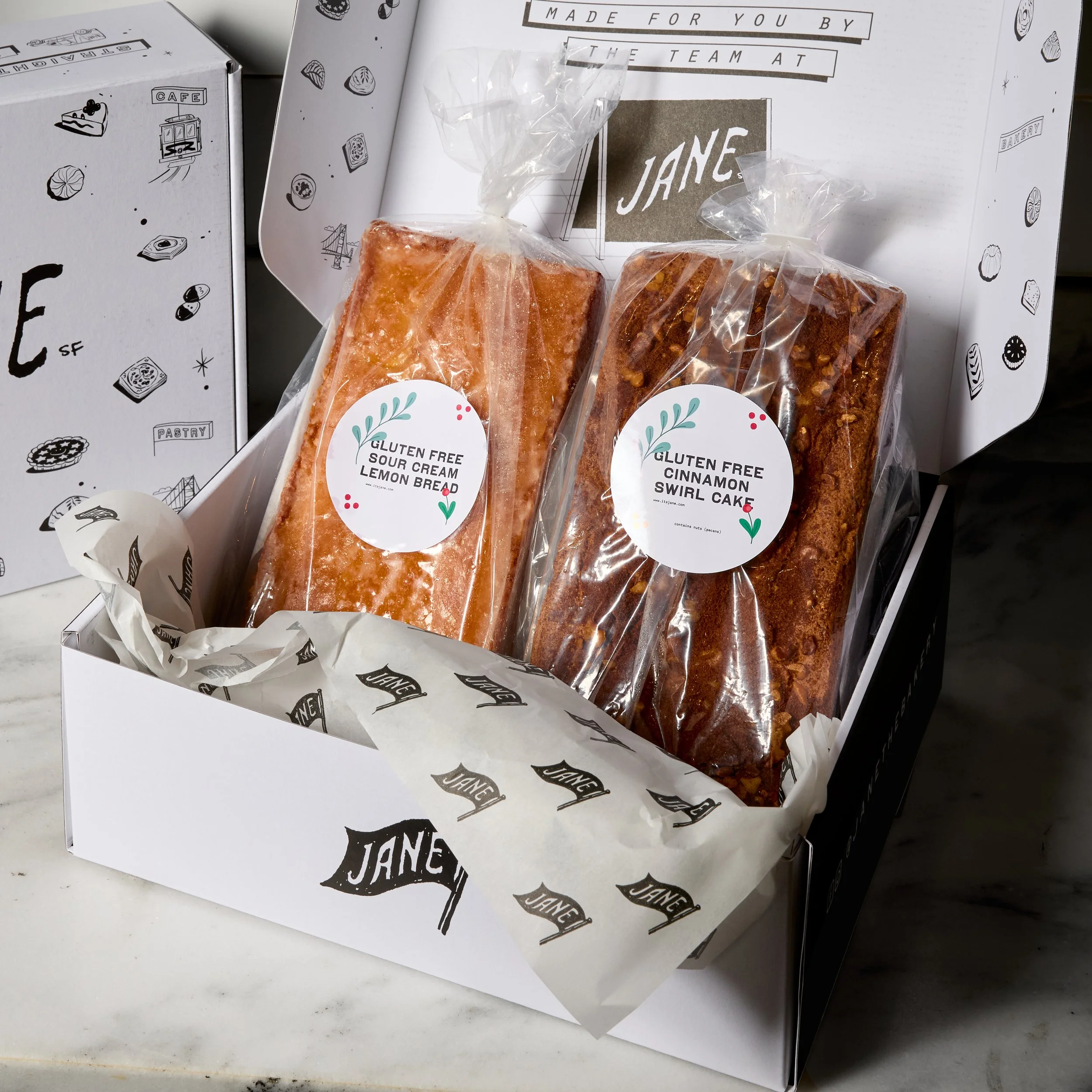A box of gluten-free baked goods containing lemon bread and cinnamon swirl cake, each wrapped in clear plastic and labeled with white stickers, placed on a white surface.