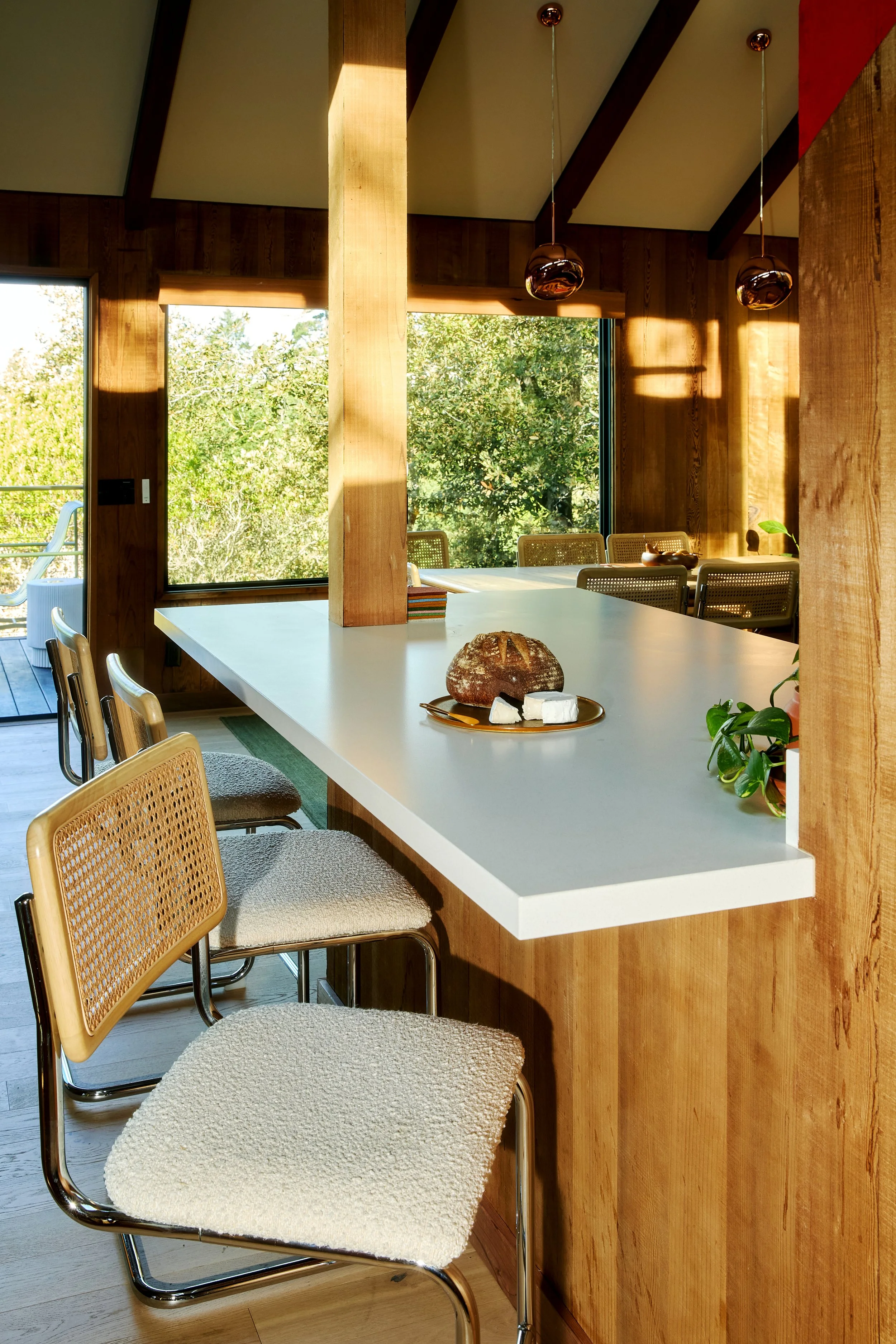 Interior of a modern kitchen or dining area with wooden walls and large windows showing greenery outside. A white countertop has a loaf of bread and cheese on a plate, with seats along the counter. Two hanging light fixtures are installed from the ce