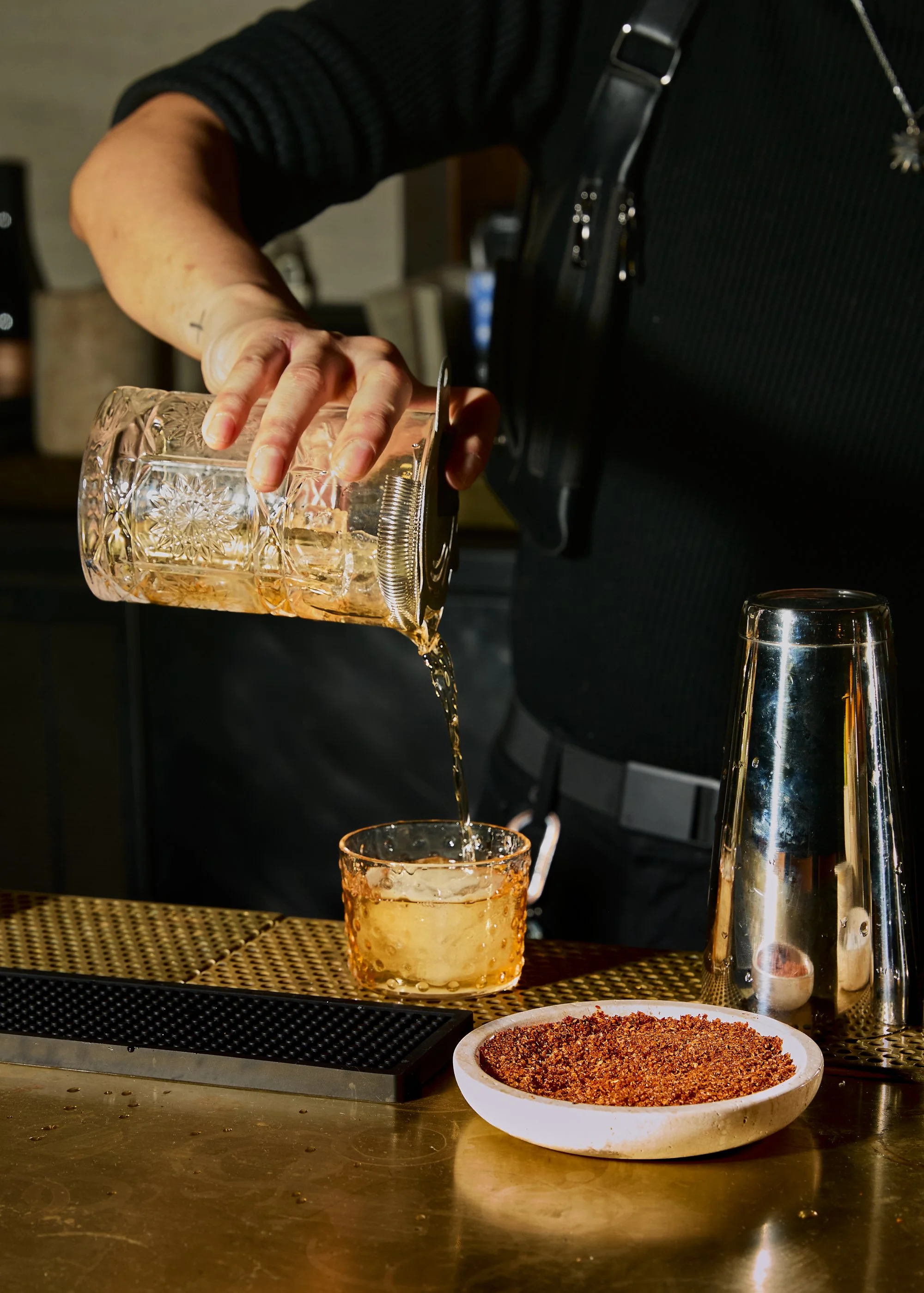 A bartender pours a drink from a glass pitcher into a short glass on a bar counter. A bowl of spice mixture and a cocktail shaker are also on the counter.