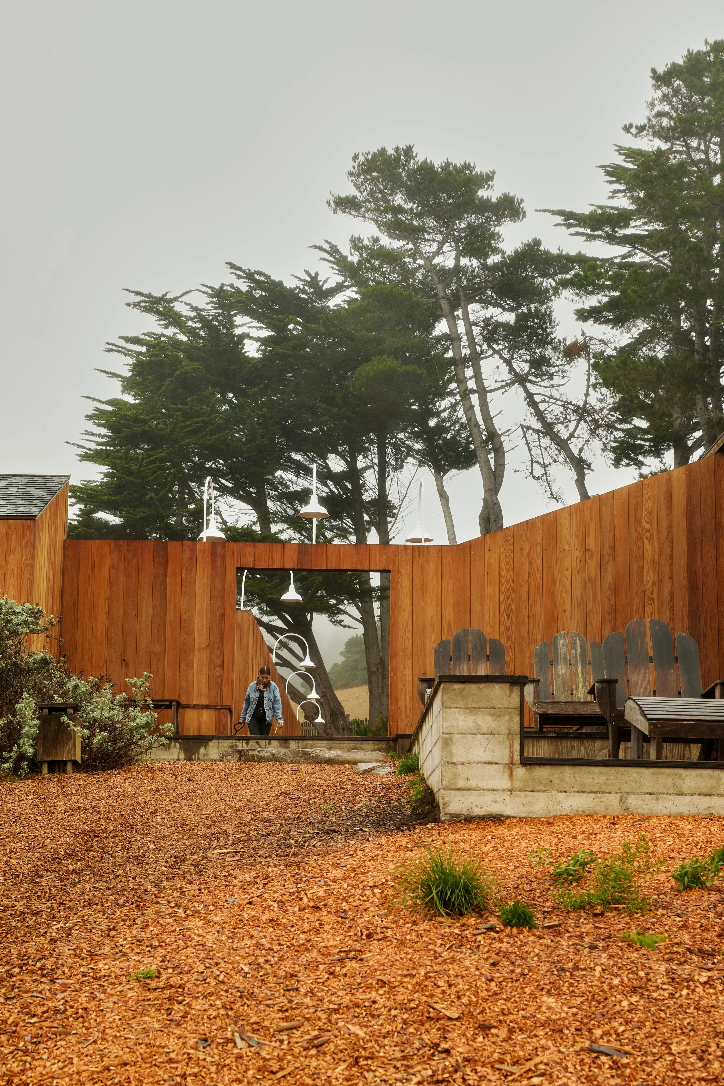 A person walking in front of a wooden structure with an opening, trees in the background, and outdoor seating on a gravel ground.