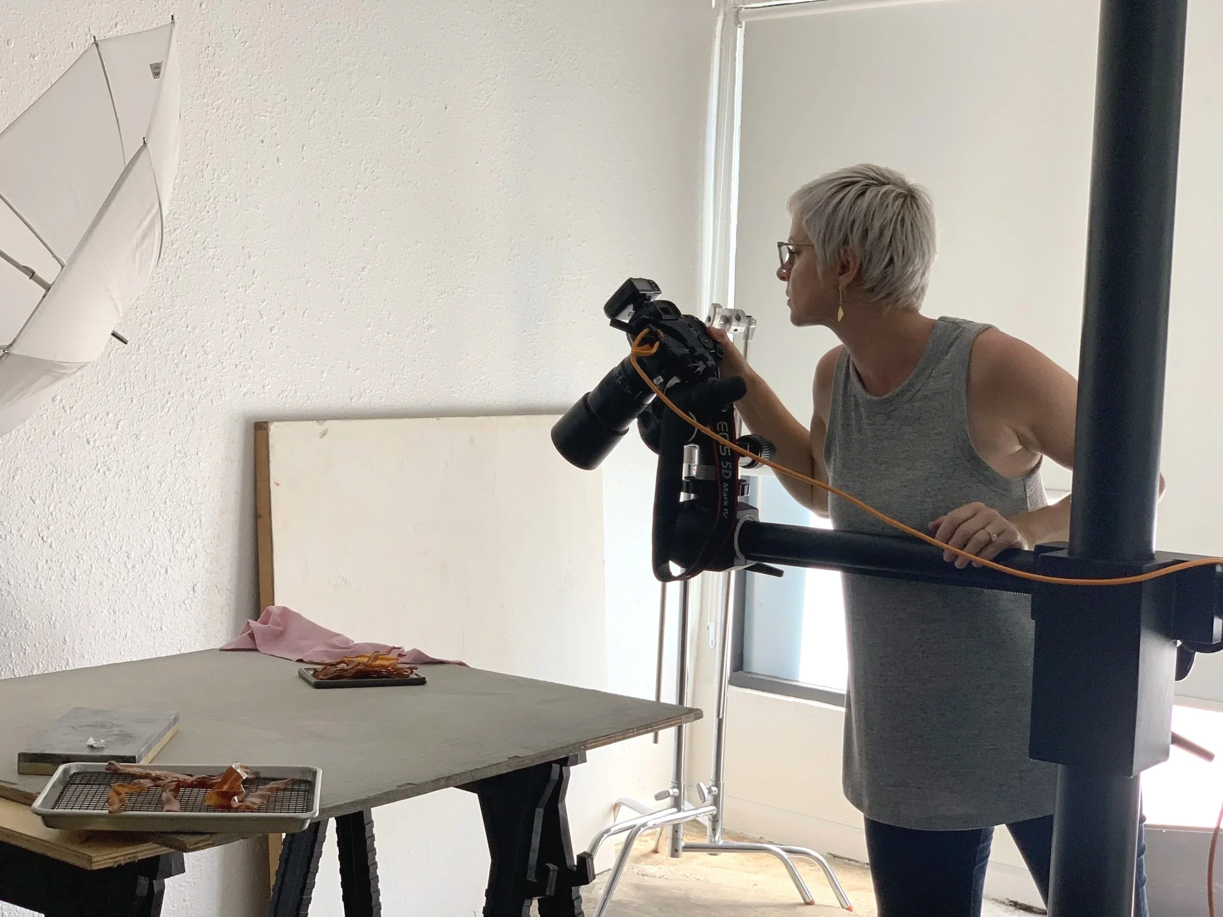 An older woman with short gray hair, glasses, and a sleeveless top, holding a camera, appears to be taking a picture or reviewing photos in a room with white walls, a table with food scraps, and a window in the background.