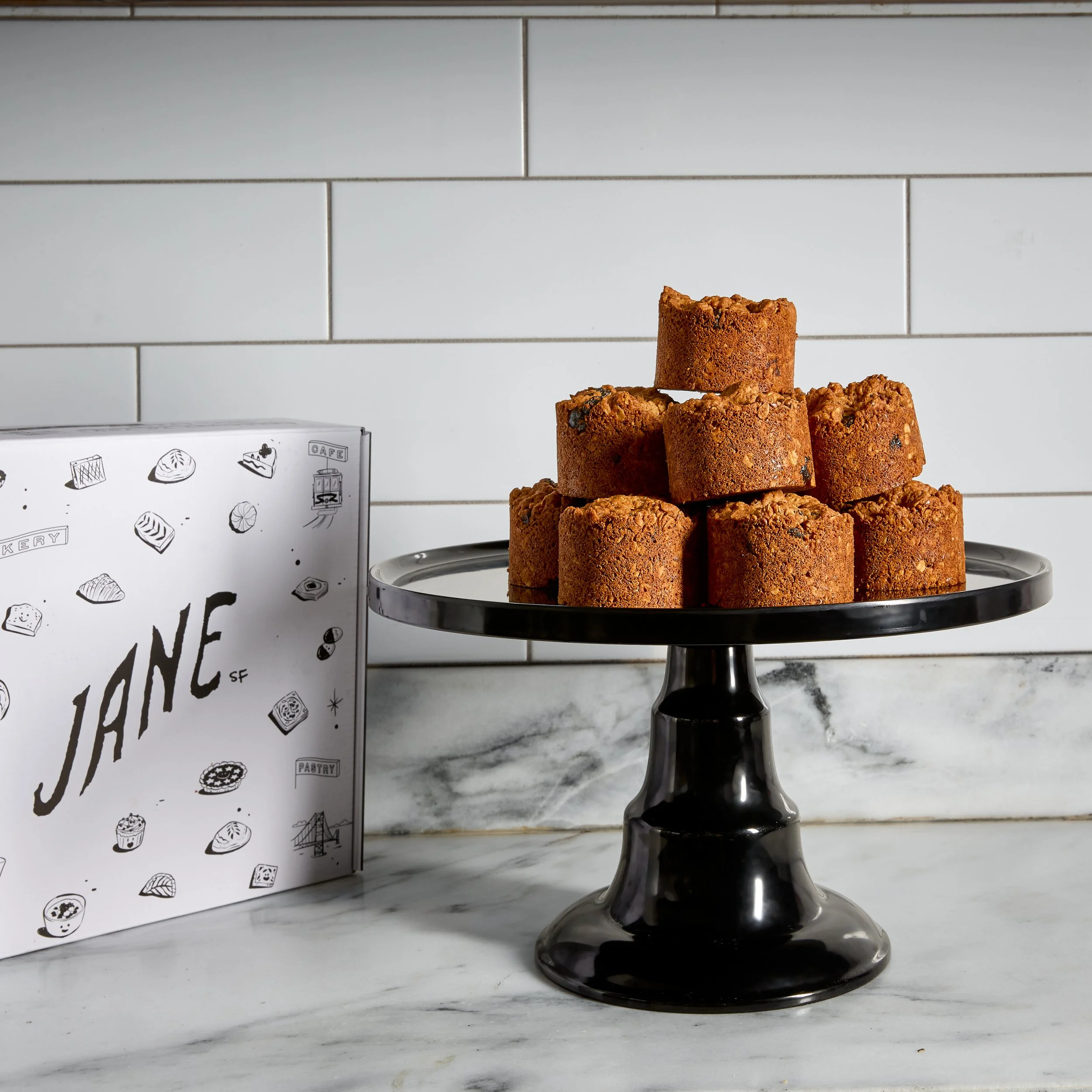 A black cake stand with a scalloped edge holding nine breaded, fried, square pieces of food, possibly croquettes, arranged in a pyramid shape. To the left, there is a white box with black line drawings of various pastries and the word "JANE SF" writt