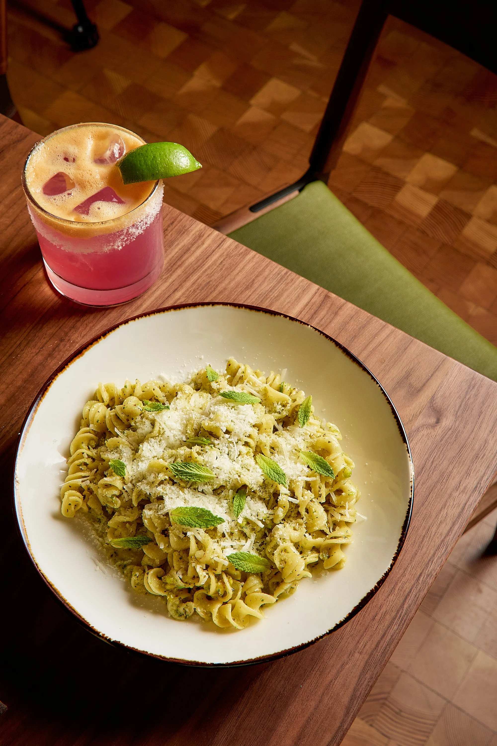 A bowl of creamy pasta garnished with fresh basil and grated cheese, with a side glass of pink cocktail with lime and ice cubes on a wooden table.