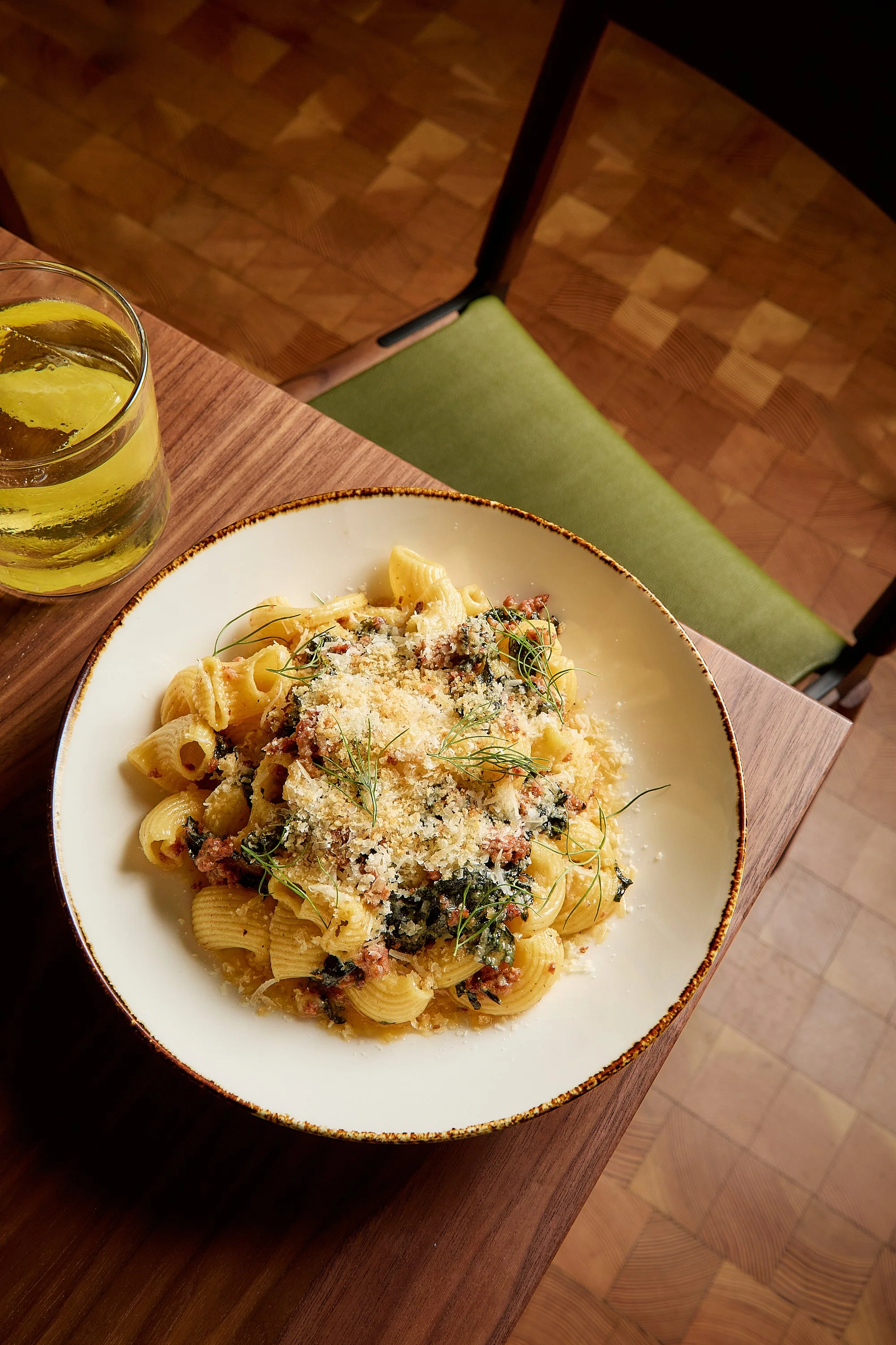 A plate of pasta with creamy sauce, sprinkled with grated cheese and garnished with herbs, next to a glass of yellow-colored beverage on a wooden table. A green cushioned chair and parquet flooring are visible in the background.