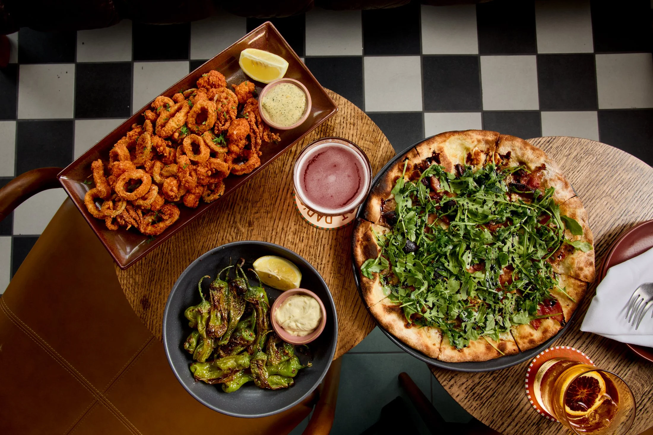 Top-down view of a table with a pizza topped with greens, a bowl of grilled peppers, a bowl of salad, fried calamari with lemon wedges, and assorted drinks.