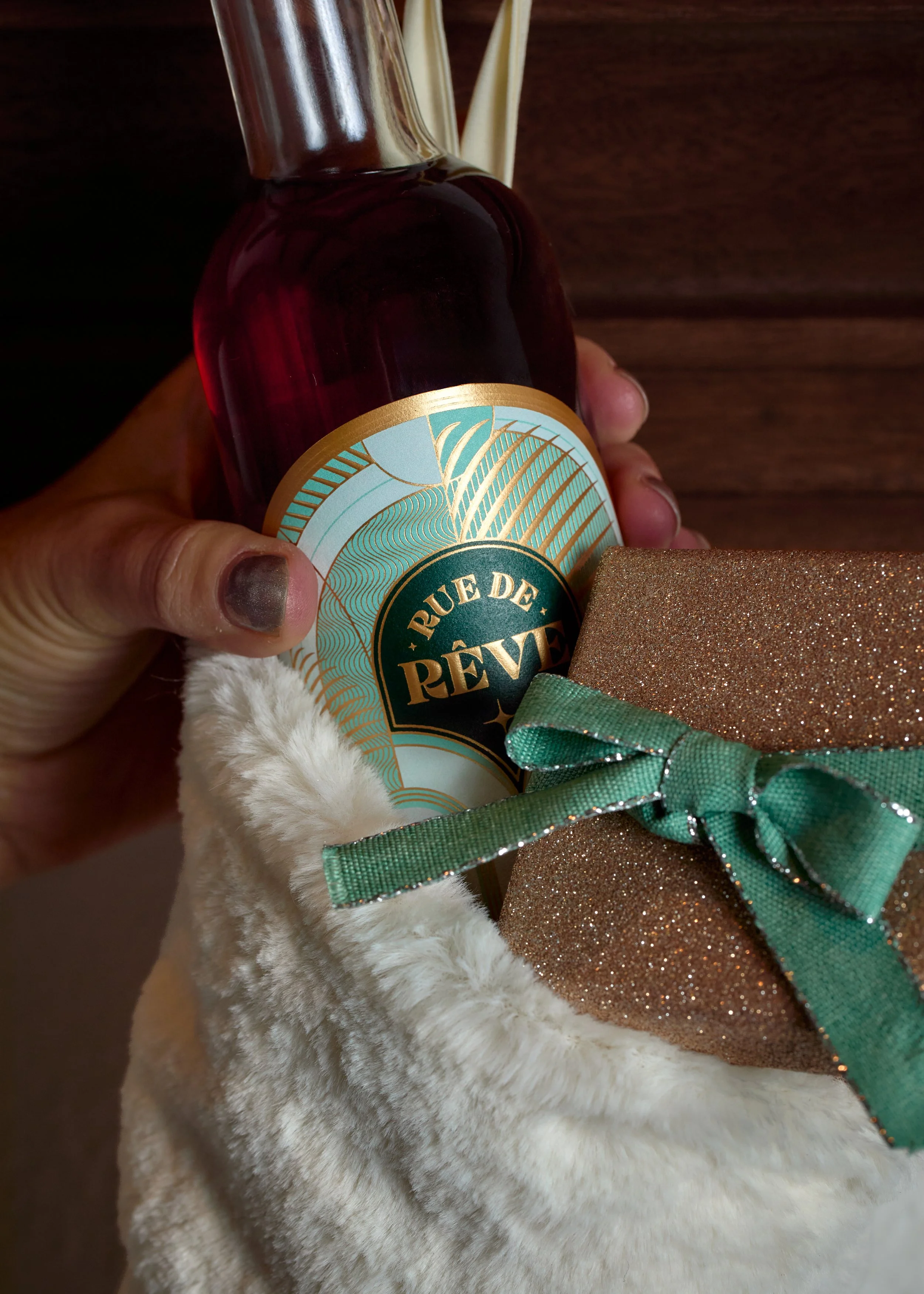 A hand holding a bottle of perfume labeled "RUE DE RÊVES" along with a small, glittery gift box wrapped with a green ribbon, all inside a plush, cream-colored pouch with a green lining.