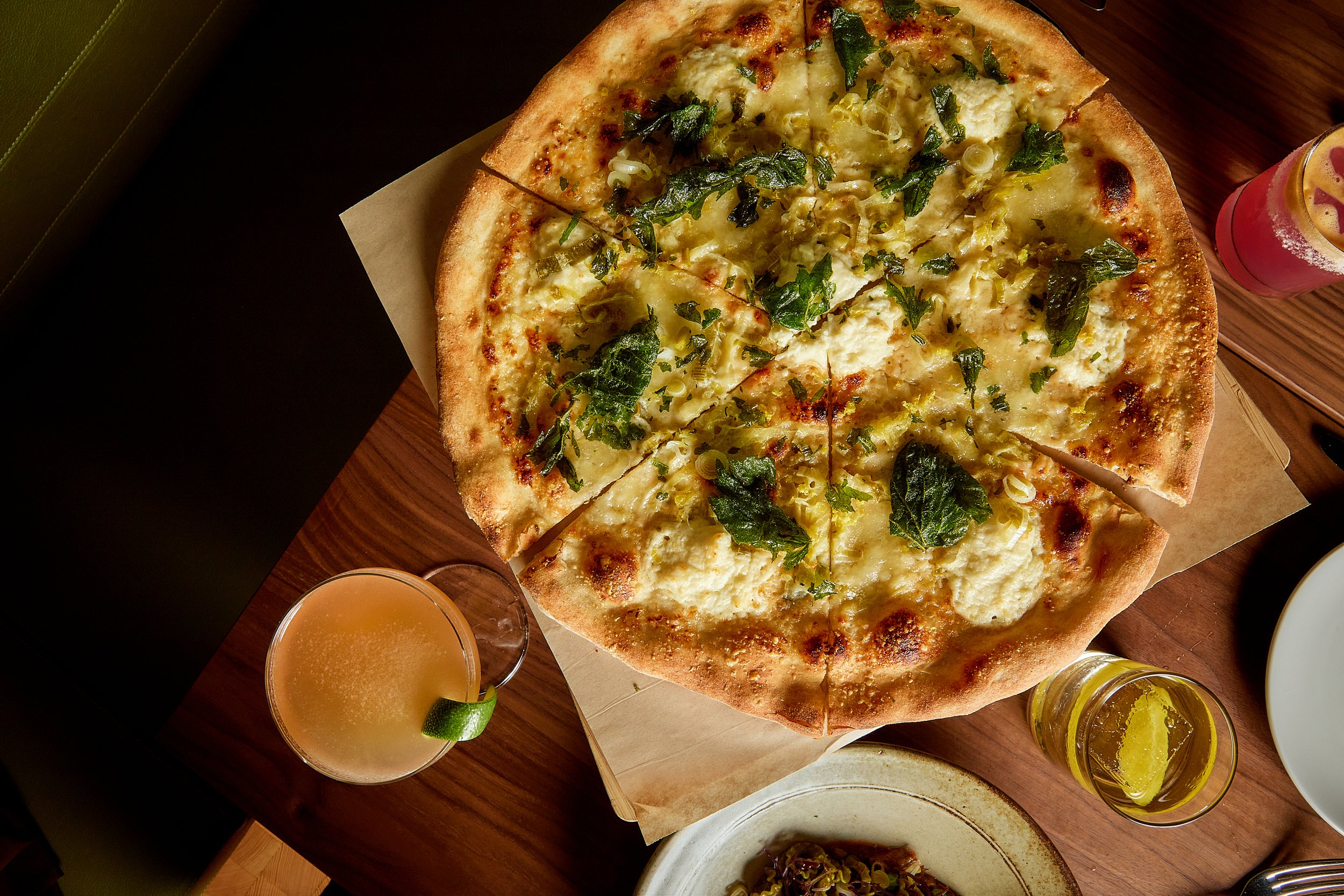A sliced pizza with toppings including cheese, green herbs, and possibly chicken on a wooden table, accompanied by glasses of orange-colored drinks with lime wedges.