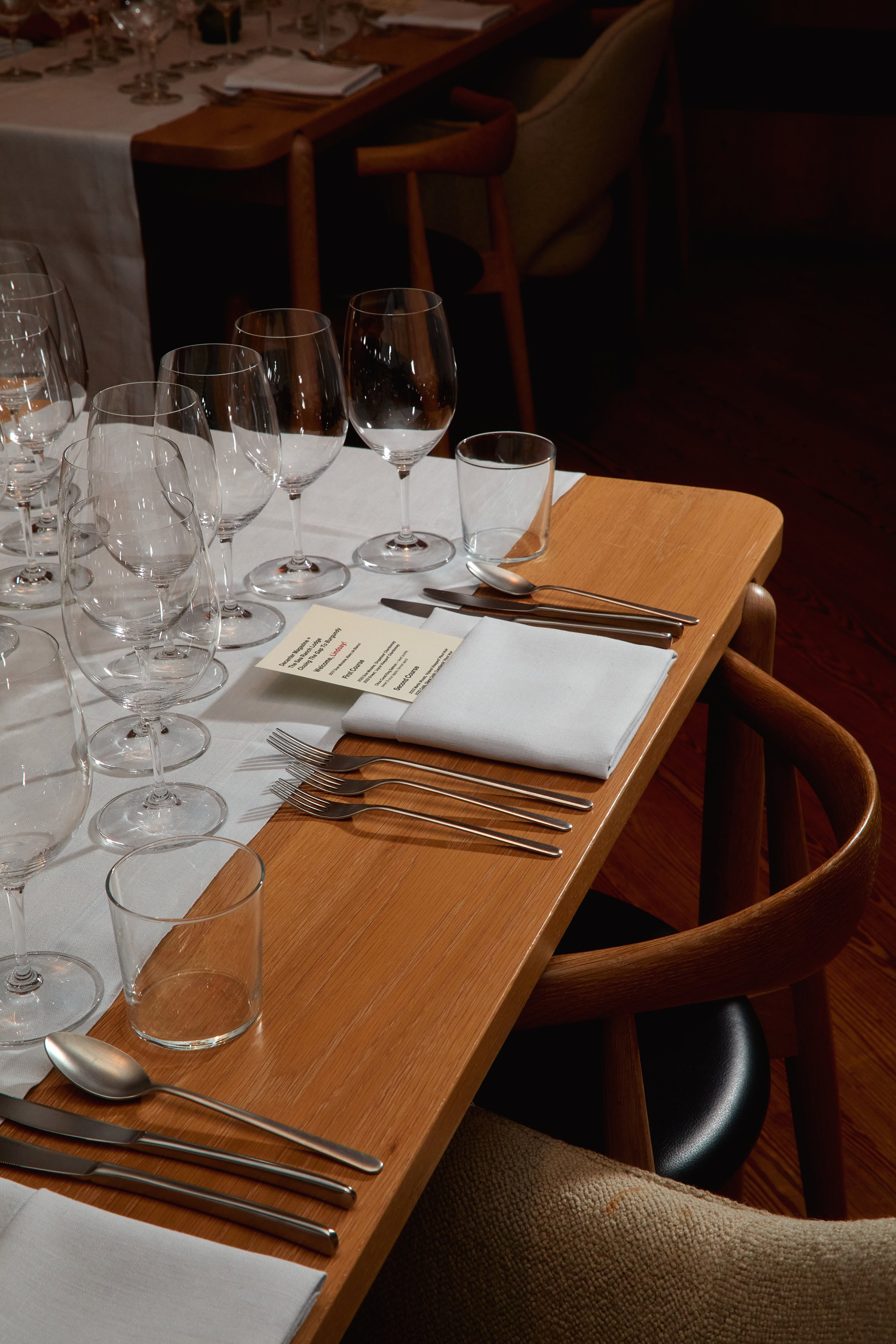 A formal dining table set with multiple wine glasses, water glasses, silverware, a white napkin, and a menu card, arranged on a wooden table with chairs around it.
