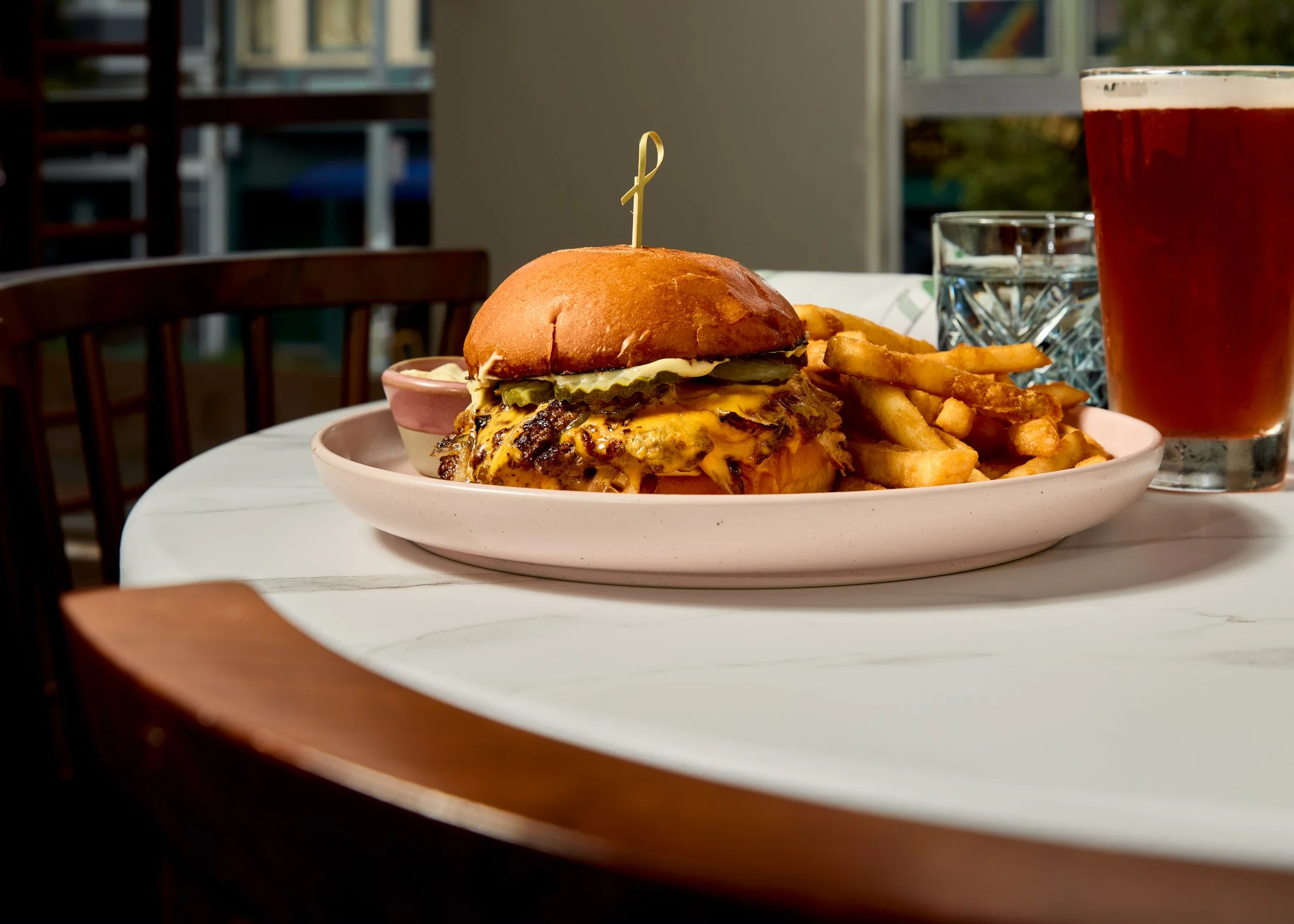A plate with a cheeseburger topped with pickles, french fries, a small pot of sauce, and a glass of soda on a white table, with a background of a balcony and trees.