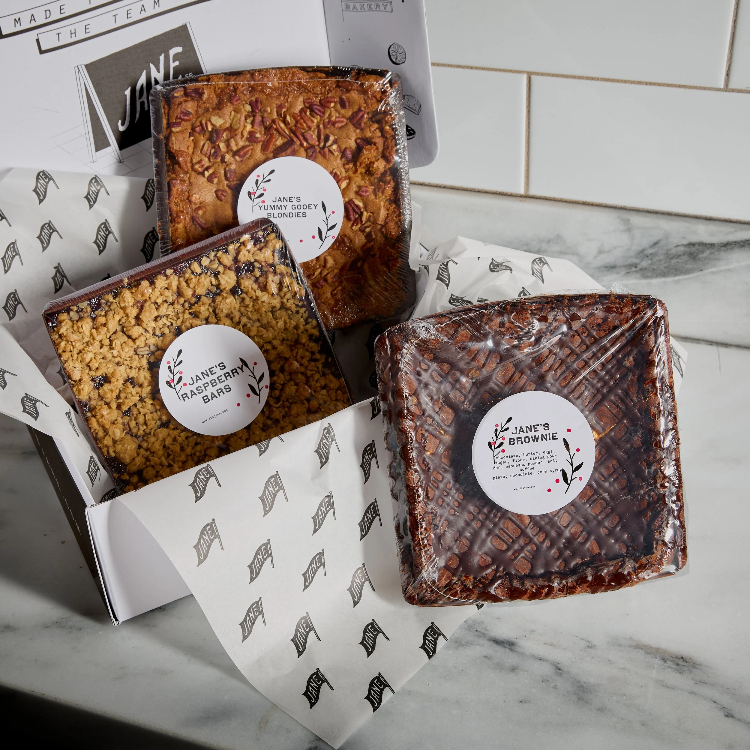 Three wrapped baked goods in a box with branded paper. The items are labeled Jane's Yummy Gooey Blondies, Jane's Raspberry Bars, and Jane's Brownie.
