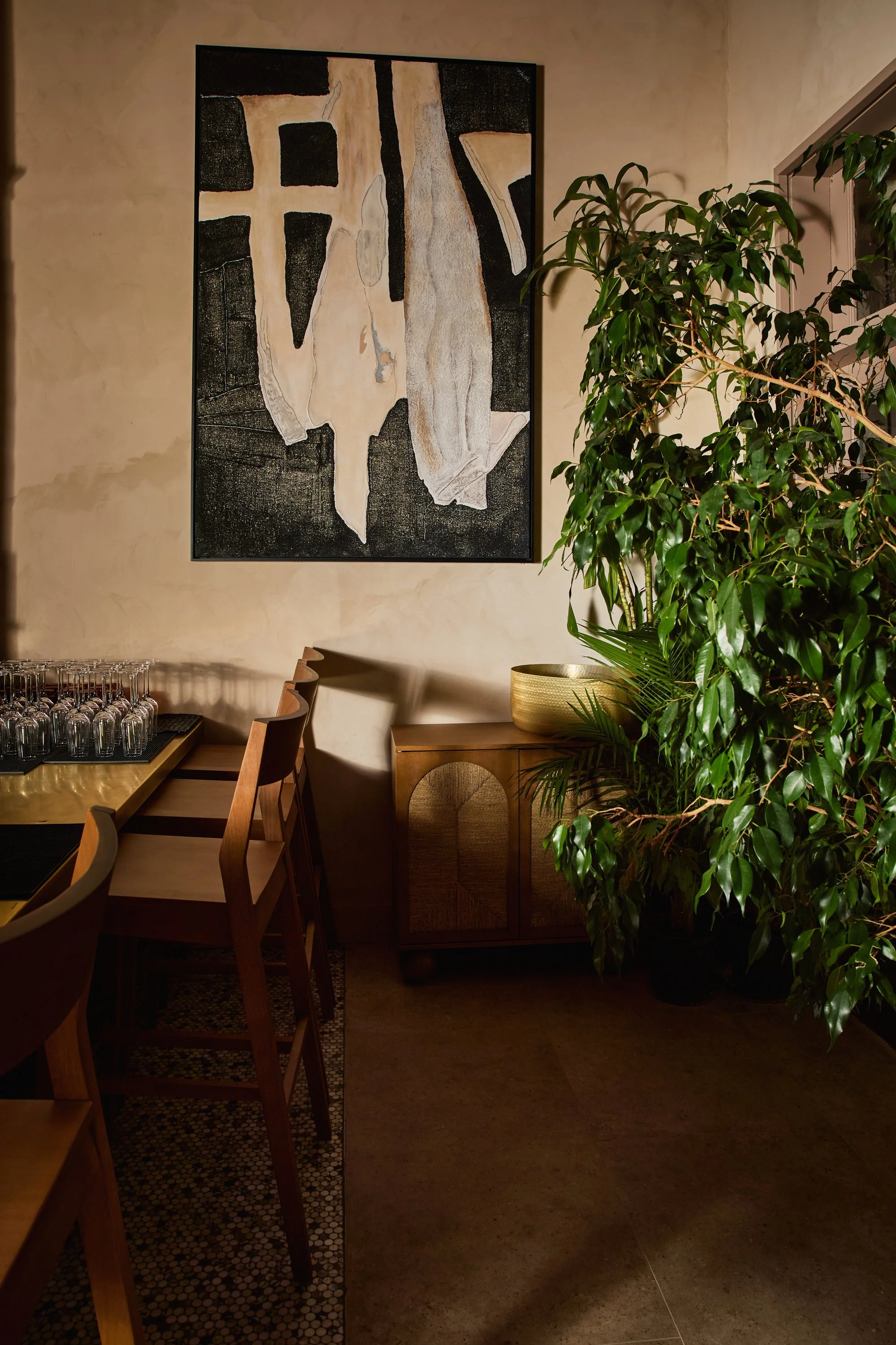 Interior view of a dining area with wooden chairs and a table set with glassware on a brass surface, a decorative wooden cabinet with an arched design, a large abstract painting on a beige wall, and a collection of lush green plants.