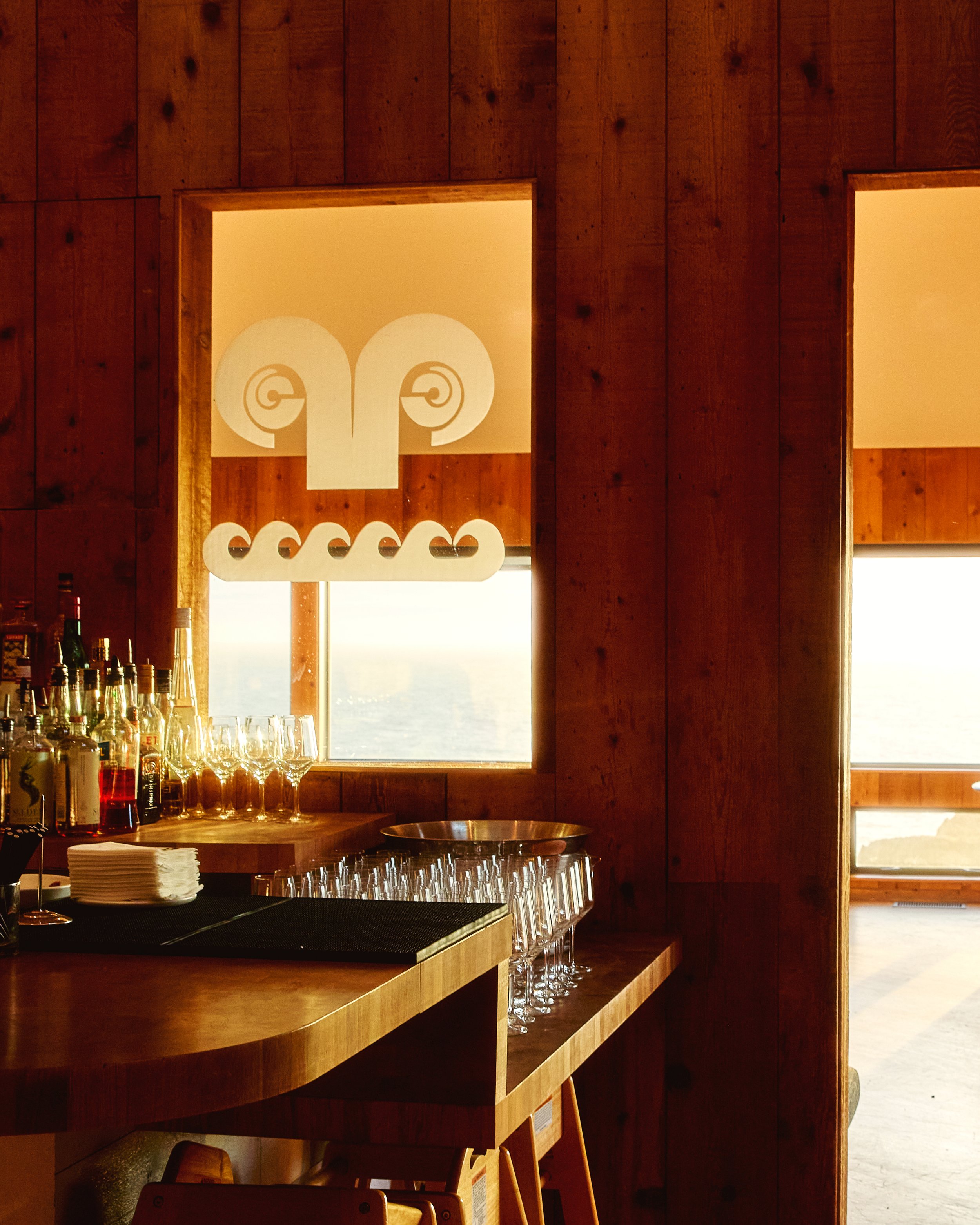 Indoor bar area with wooden walls, a window, glassware, and bottles, featuring decorative art with two stylized face profiles in the window.