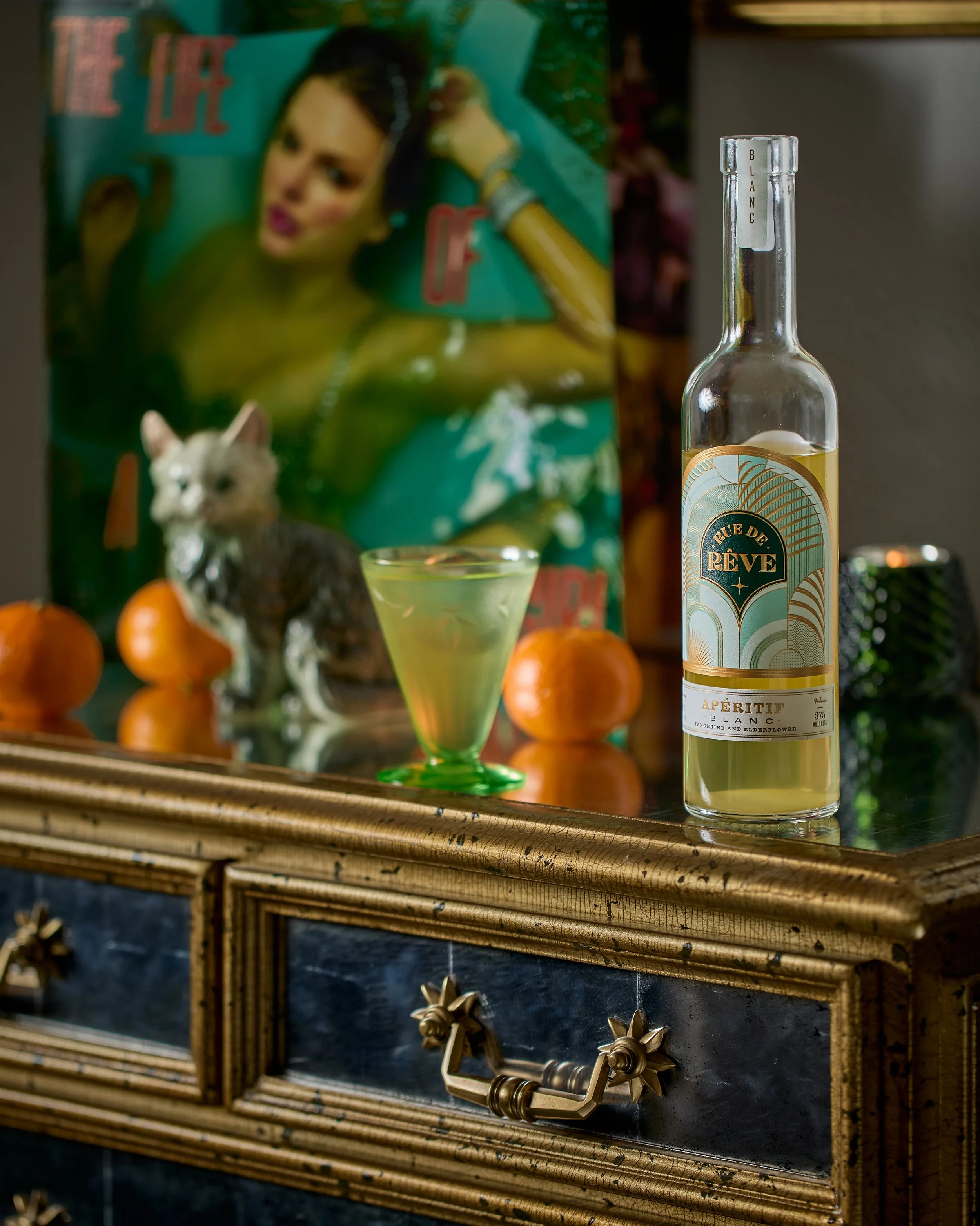 A bottle of Rue de Rêve Blanc Aperitif, a green cocktail in a glass, and an artwork featuring a woman with dark hair, along with a small figurine of a cat and oranges on a wooden surface.