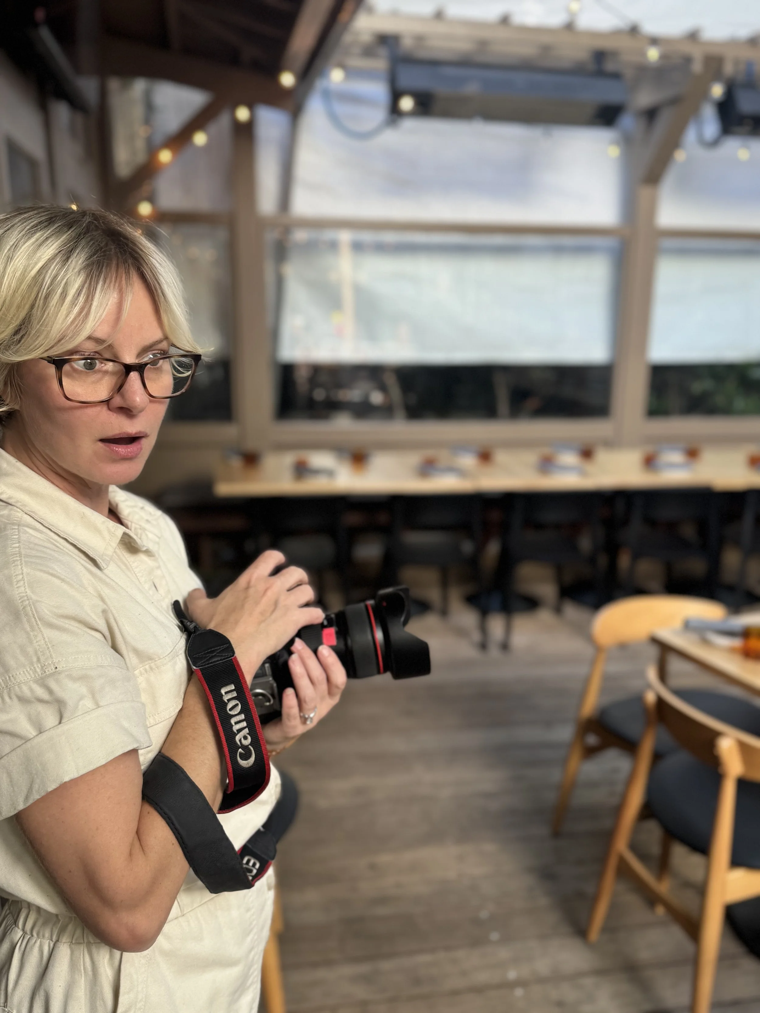 A woman with glasses holding a camera, standing indoors with a background of wooden chairs and tables, and a glass window wall showing an outdoor view.