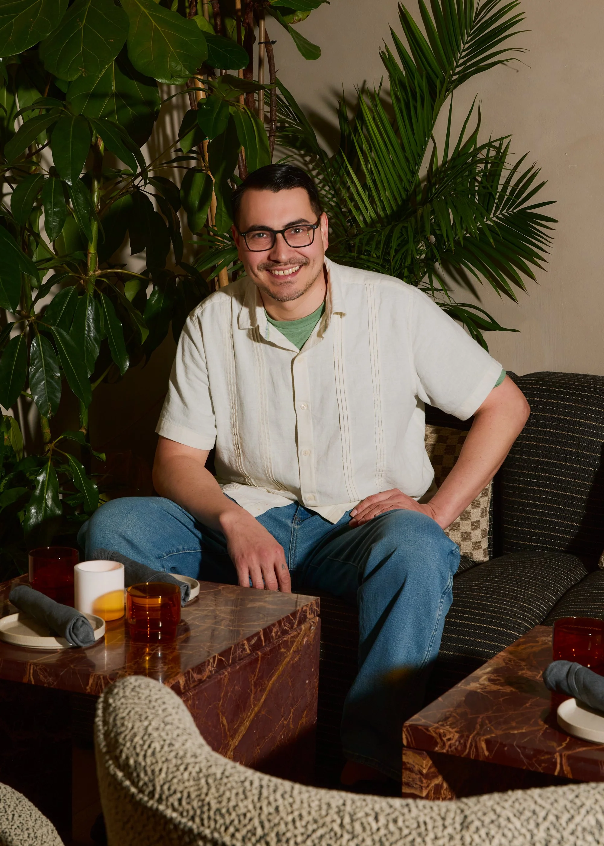 A smiling man wearing glasses, a short-sleeved white shirt, and jeans sitting on a sofa with large green plants in the background.