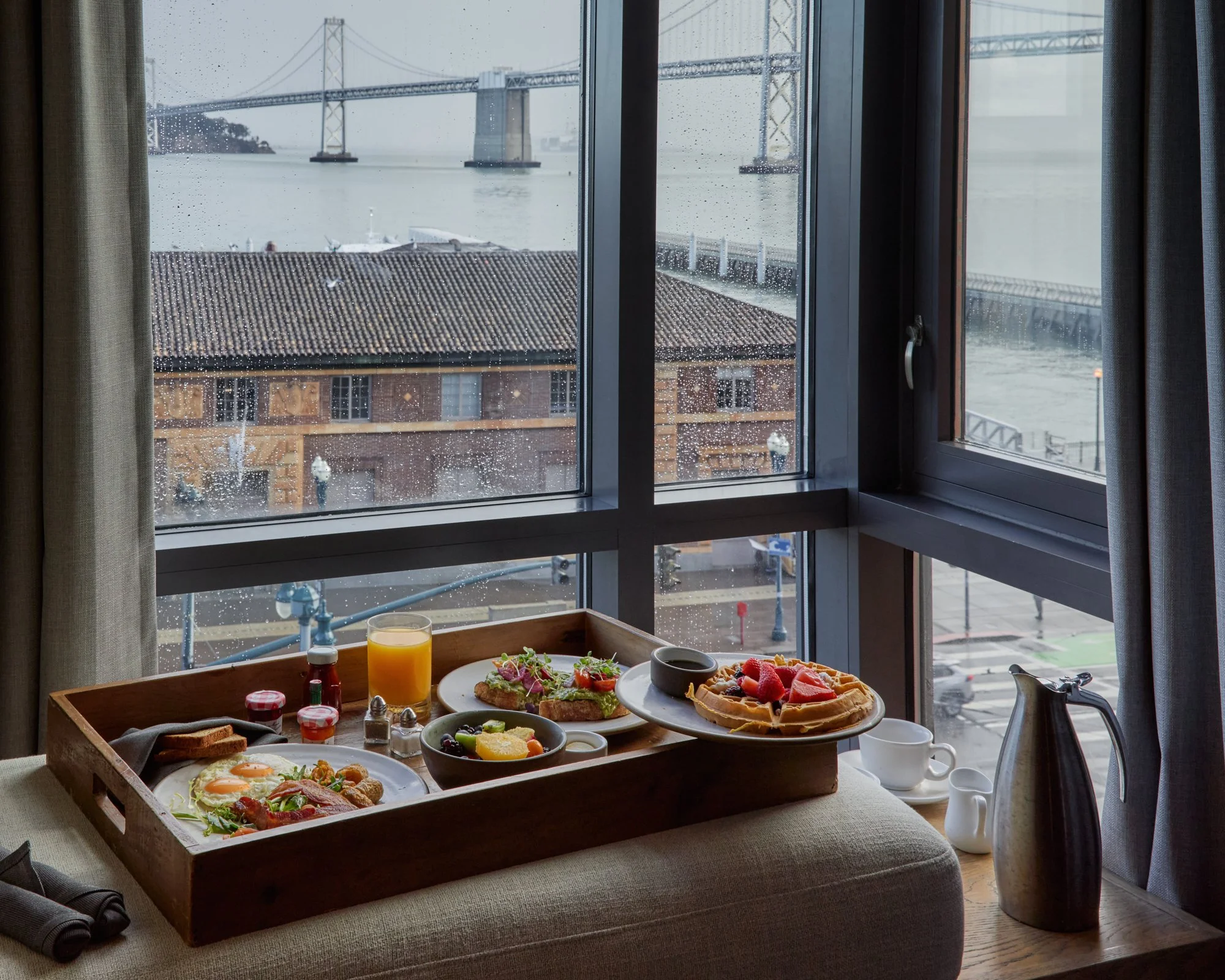 Breakfast table with eggs, toast, salad, fruit, waffles, buttermilk in a mug, juice, and condiments, near a window overlooking a rainy city with a bridge in the background.