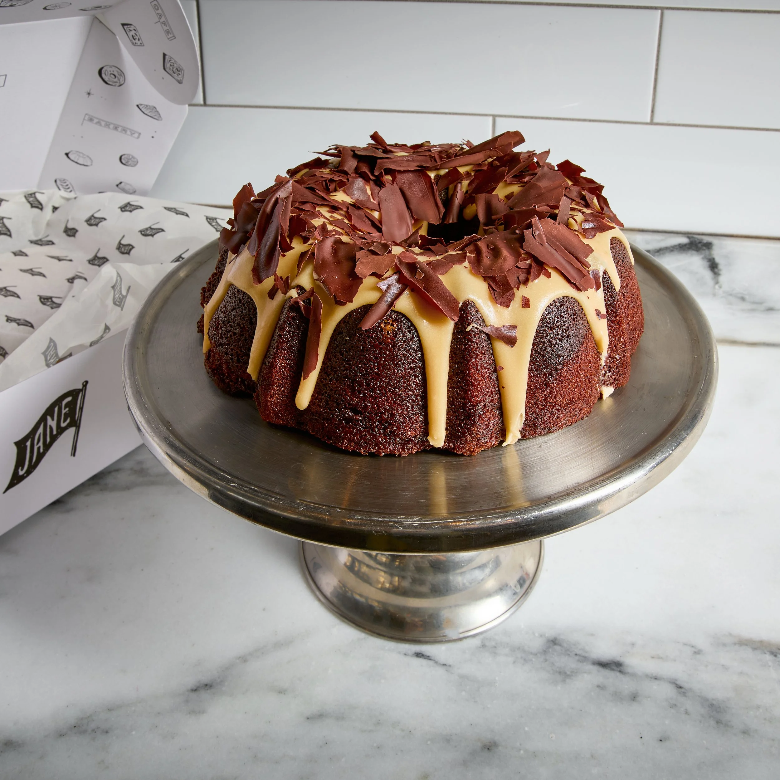 A bundt cake with chocolate shavings and white icing drizzle on a silver cake stand.