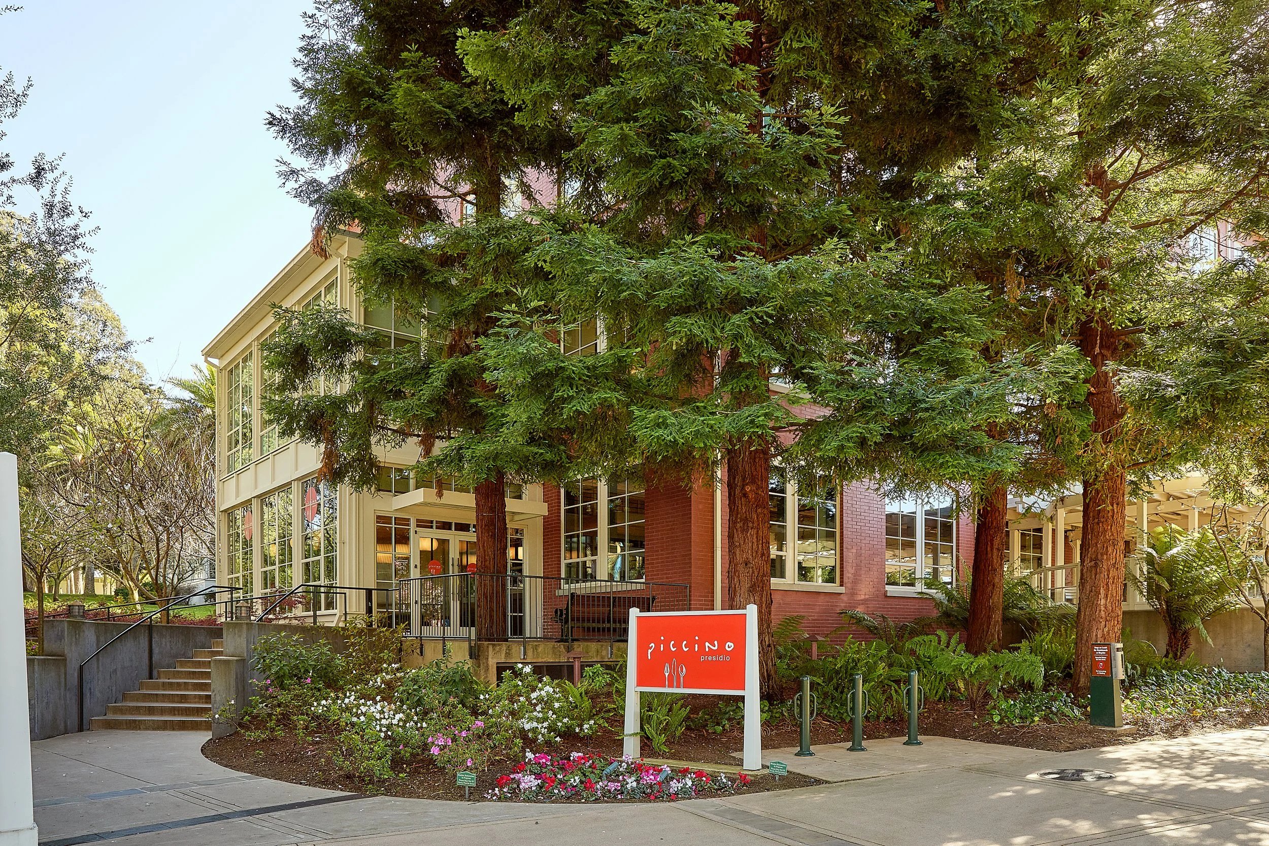 A street view of a modern building surrounded by tall evergreen trees, with a red sign that says "Piccino presido" and flowers in the foreground.